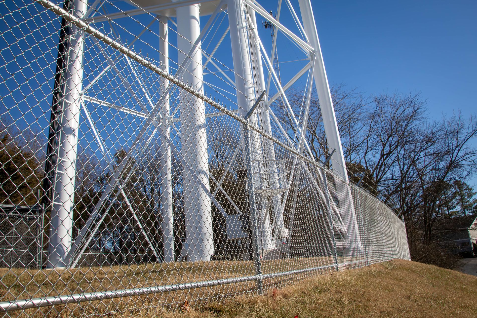 Chain-link fence surrounding a tall, white water tower, under a clear blue sky.
