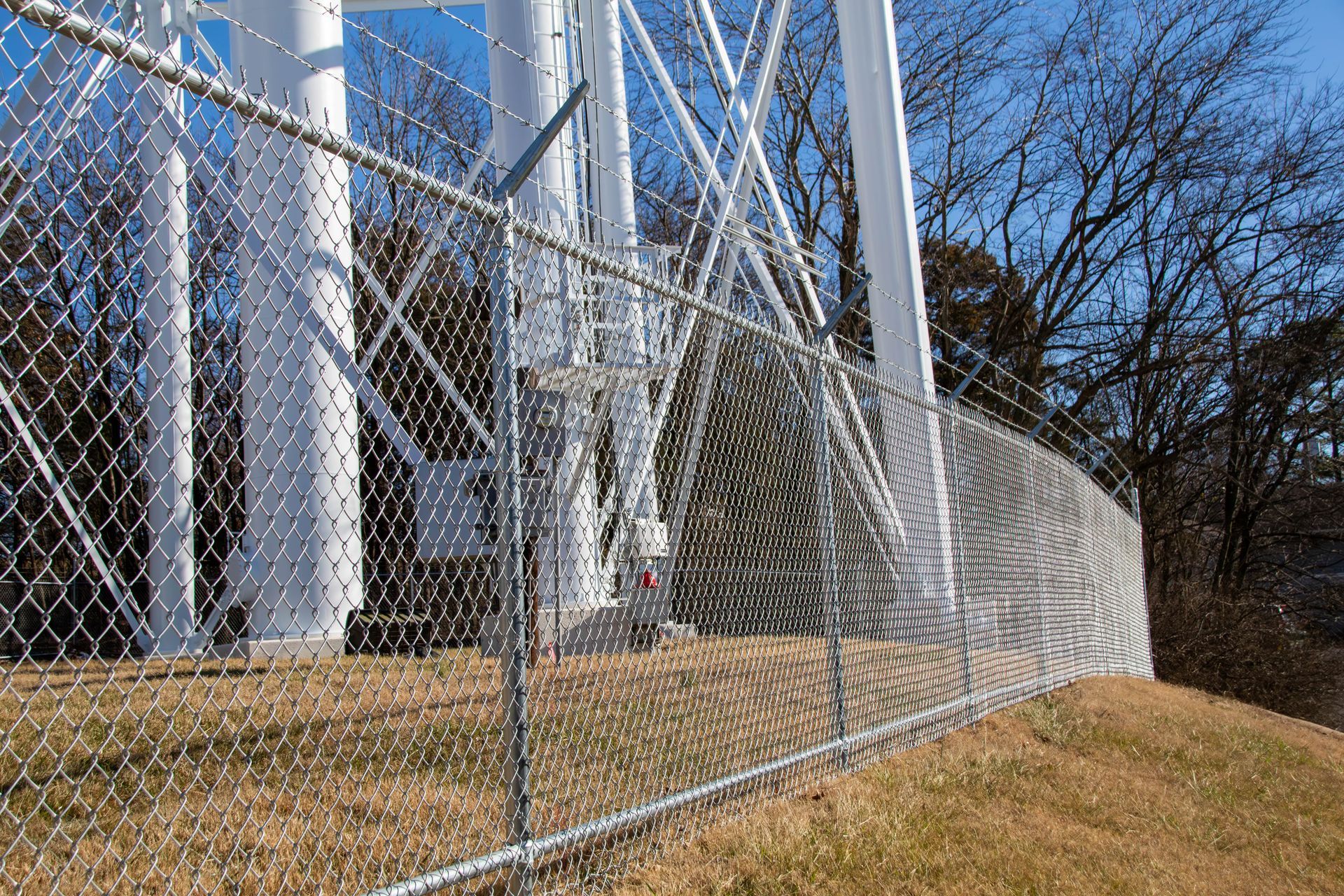 Chain-link fence surrounding a white communication tower on a hill with brown grass, trees, and blue sky.
