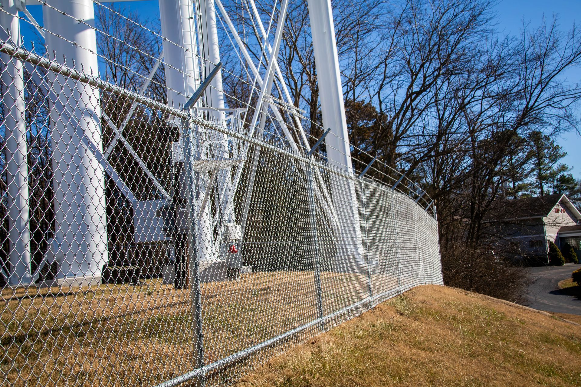 Chain-link fence surrounding a white water tower, on a grassy hill, with trees and a house in the background.