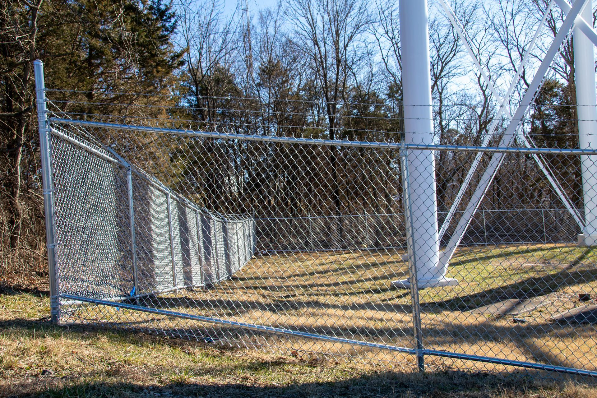 Chain-link fence surrounding a white tower structure, set in a grassy area with trees and blue sky.