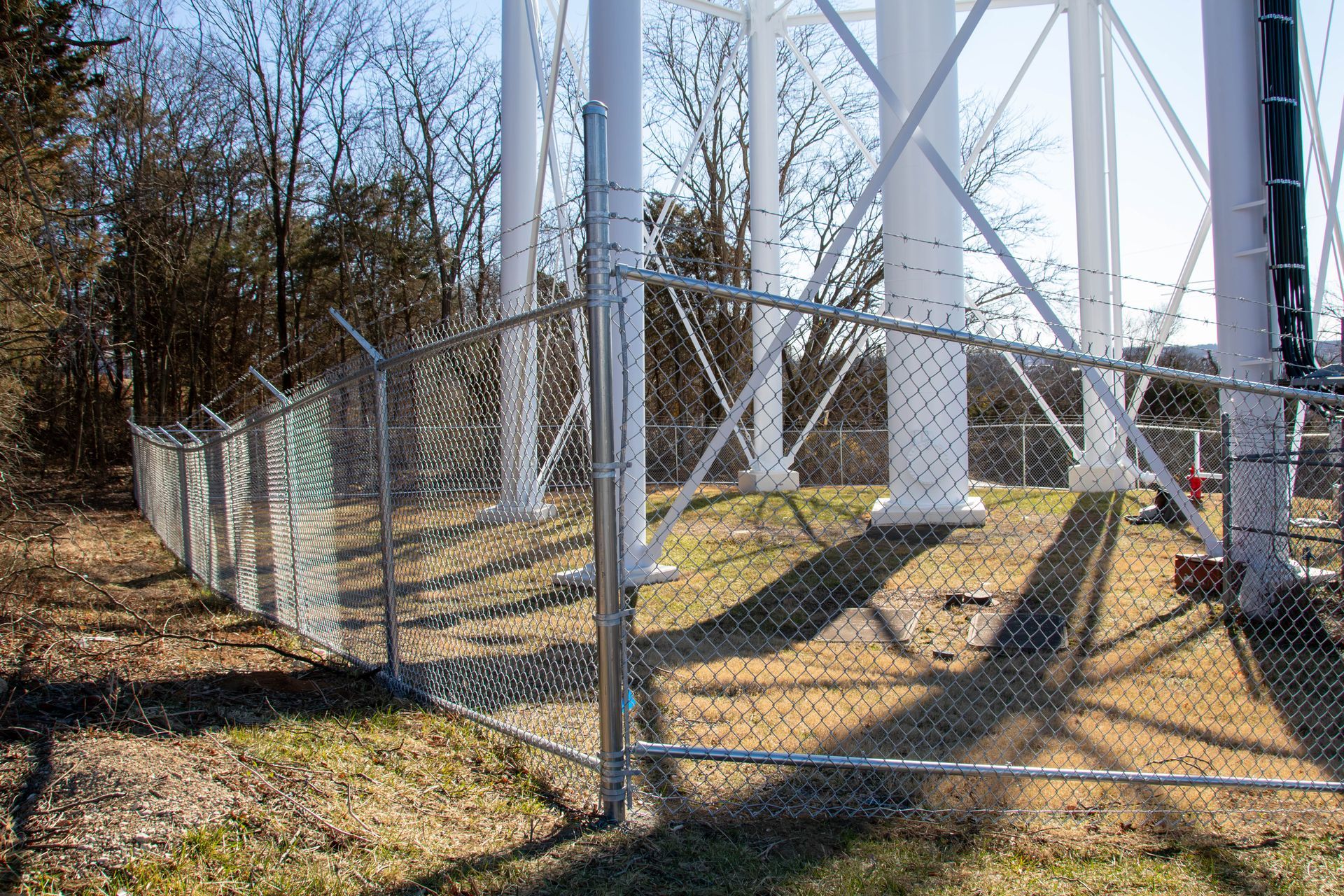Chain-link fence with barbed wire surrounds a white water tower structure in a grassy, wooded area.