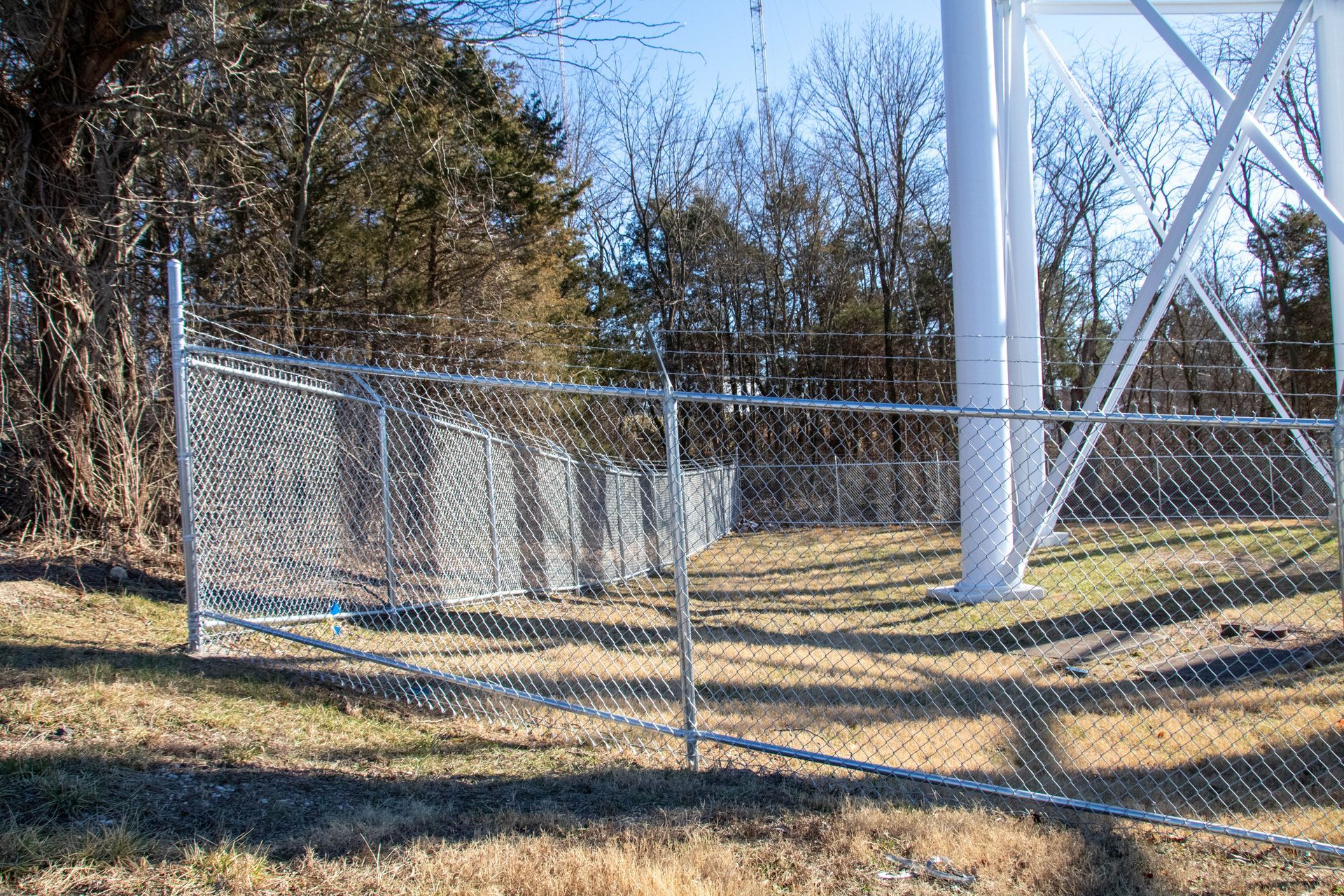 Chain-link fence surrounding a water tower, with trees in the background under a blue sky.
