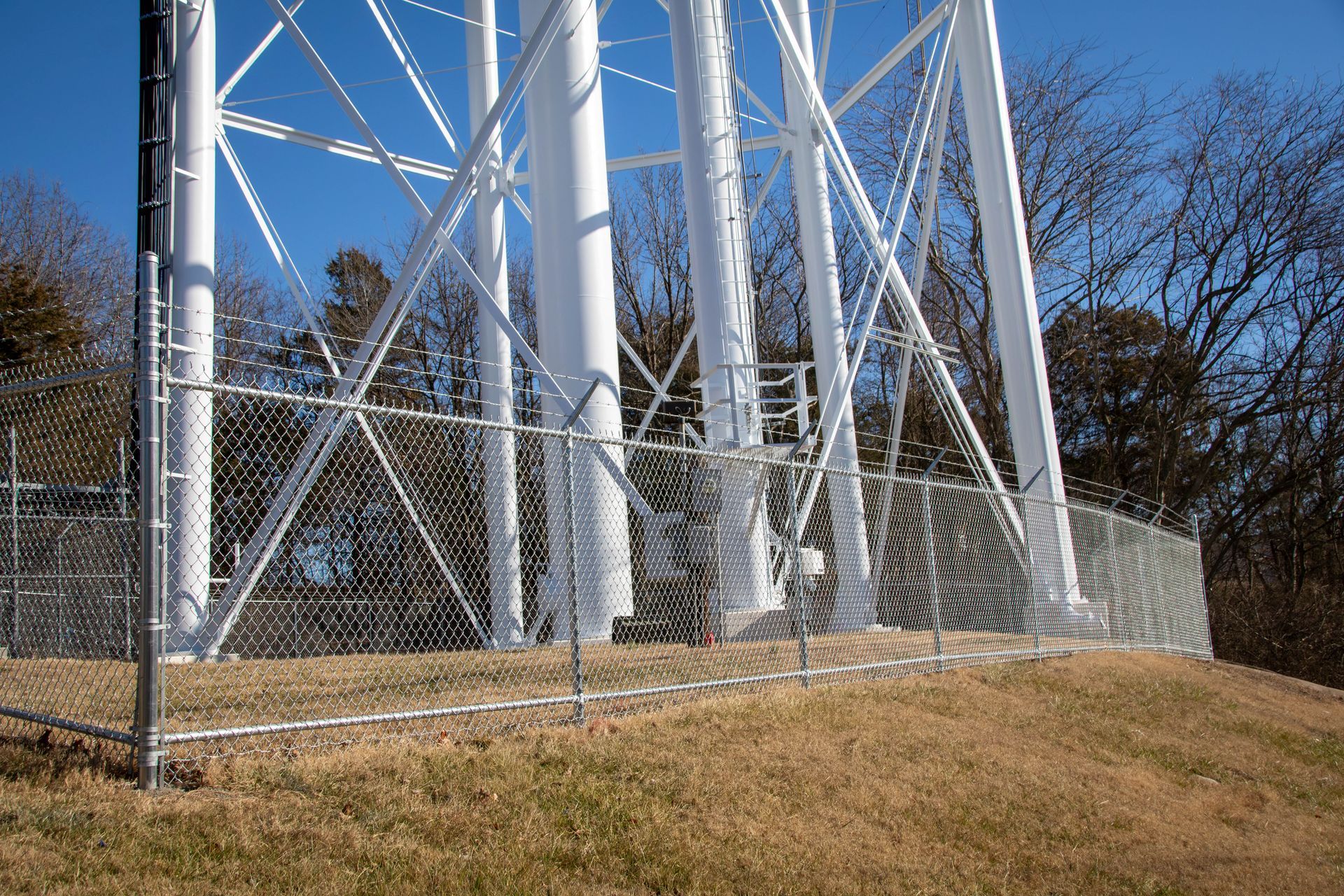 White communication tower base surrounded by a chain link fence on a grassy hill, blue sky background.