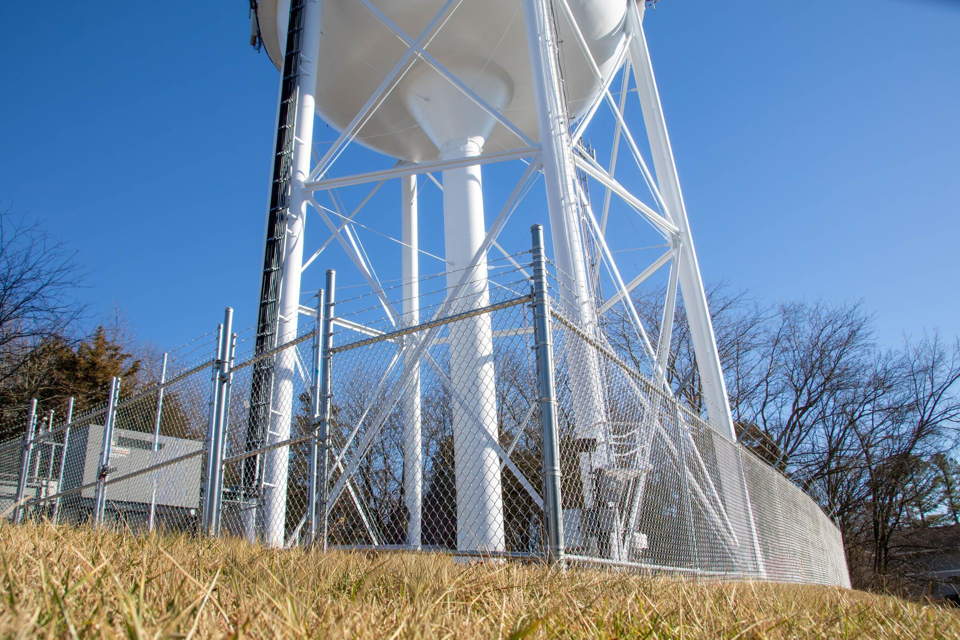 White water tower with chain-link fence on a grassy hill, against a blue sky.