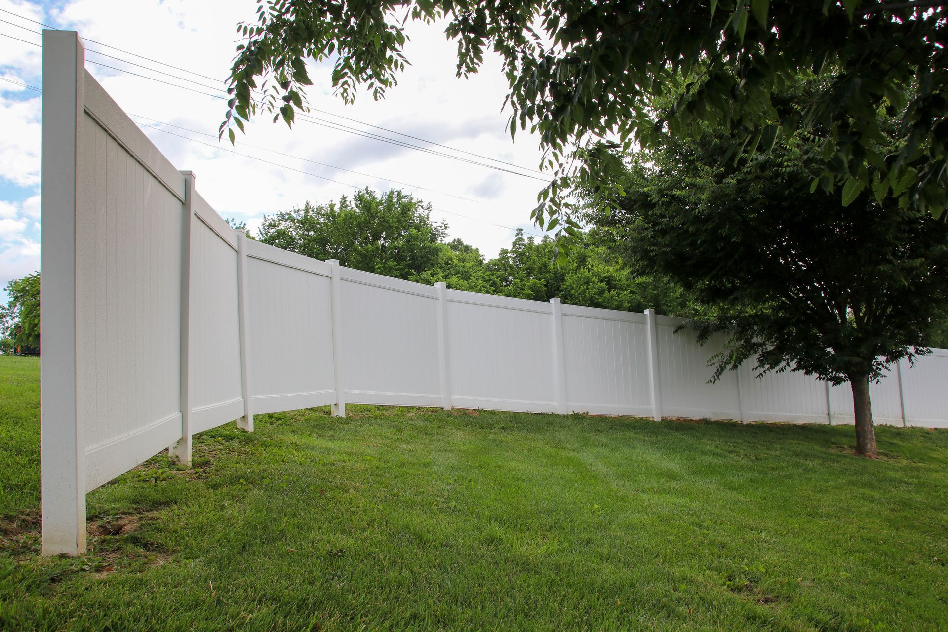 White vinyl fence curving across a grassy yard, trees in the background, blue sky with clouds.