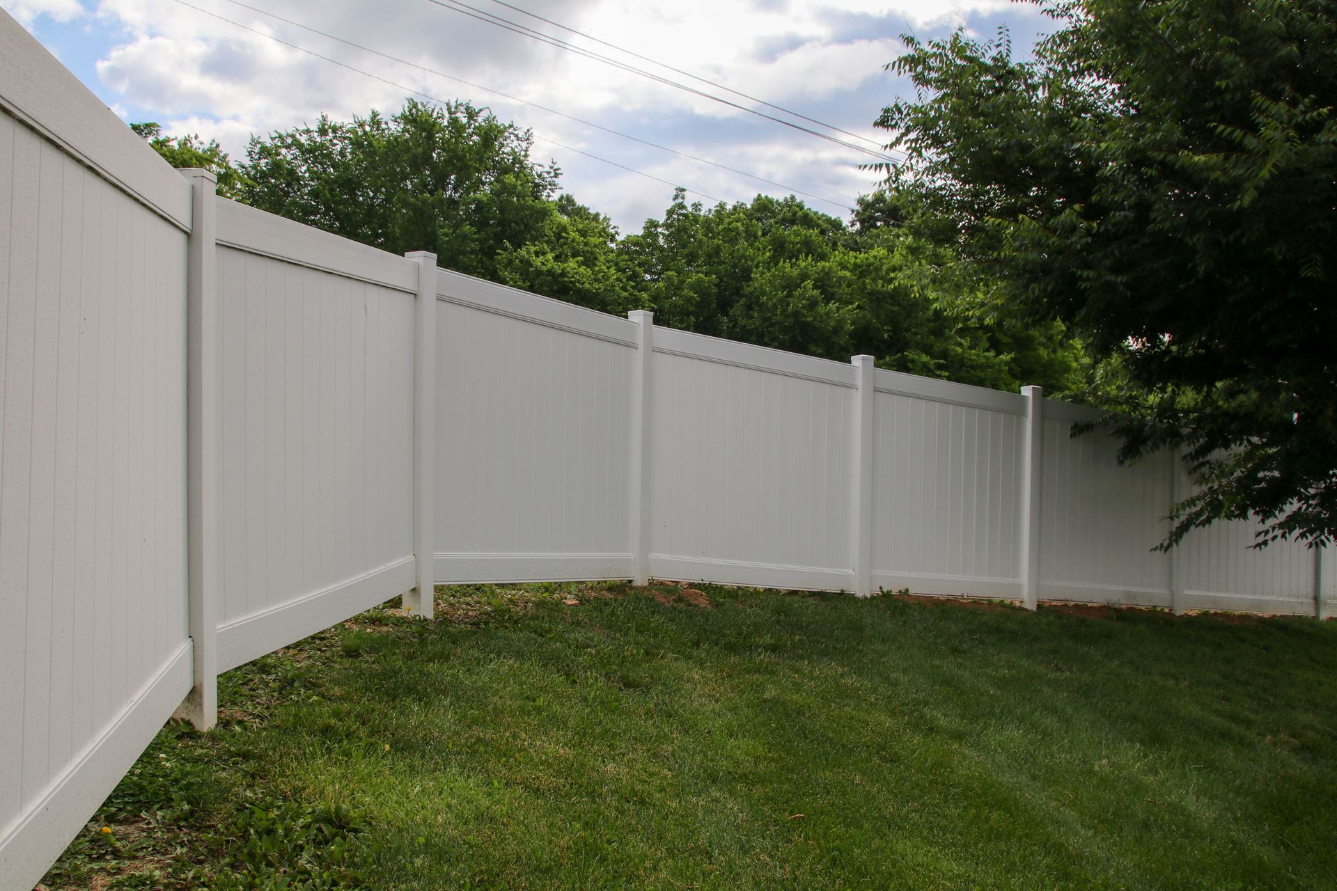 White vinyl privacy fence in a grassy yard, with trees and a cloudy sky in the background.