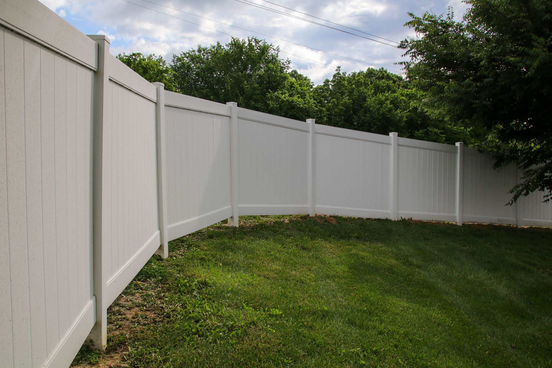 White vinyl fence surrounding a grassy yard with trees in the background under a cloudy sky.