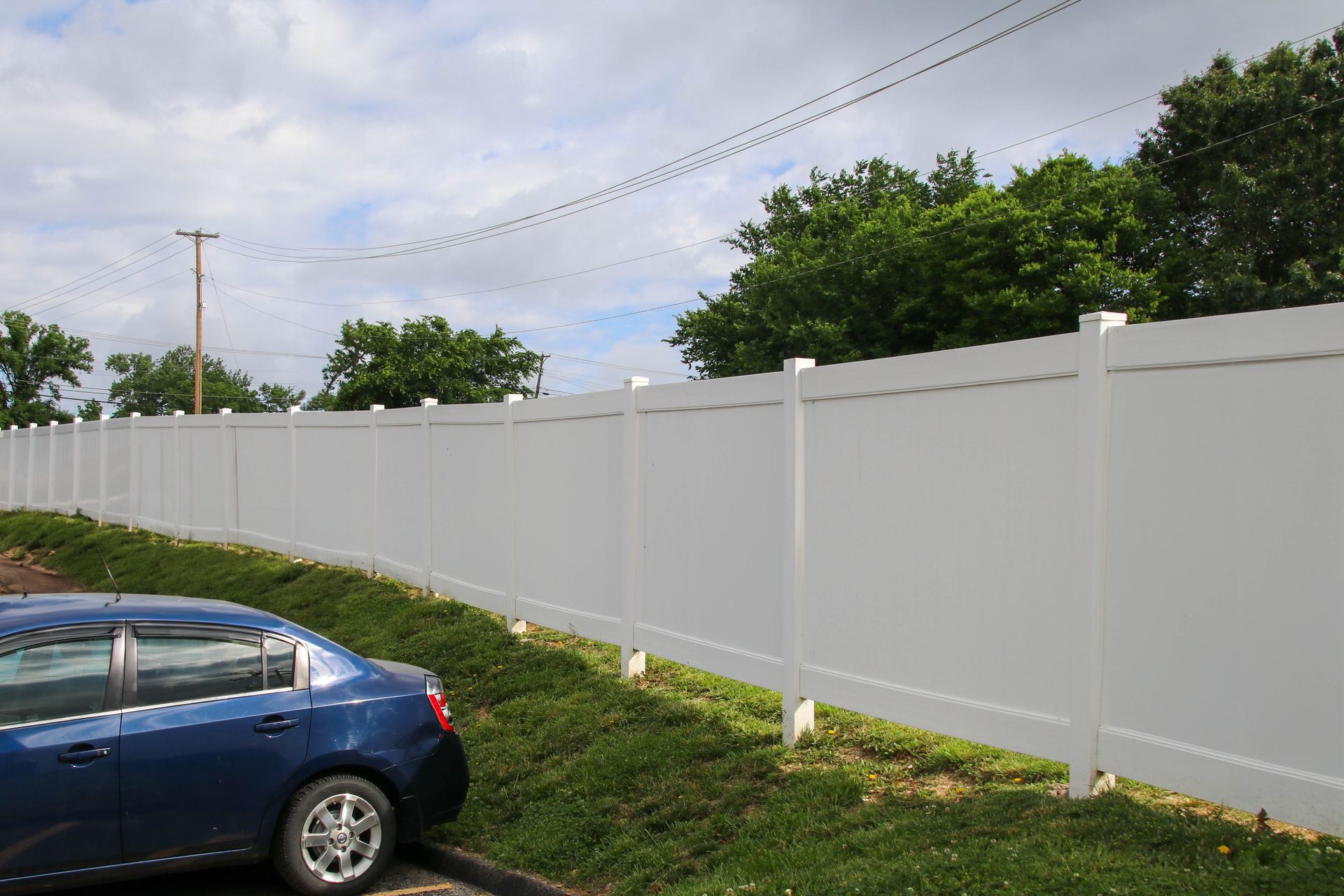Blue car parked next to a long, white vinyl fence on a grassy hill; trees and a cloudy sky in the background.