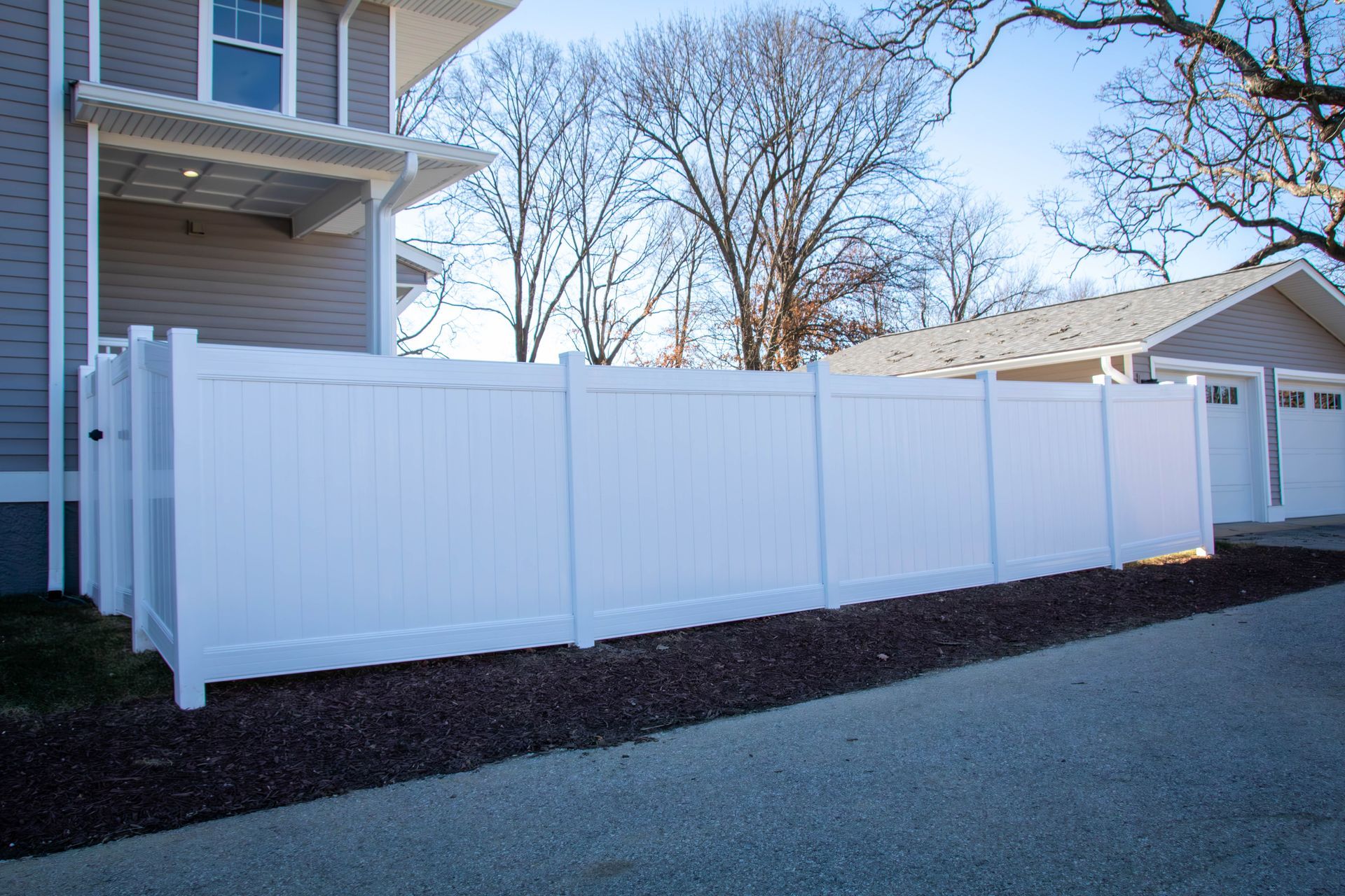 White vinyl fence along the side of a house and garage; blue sky overhead.