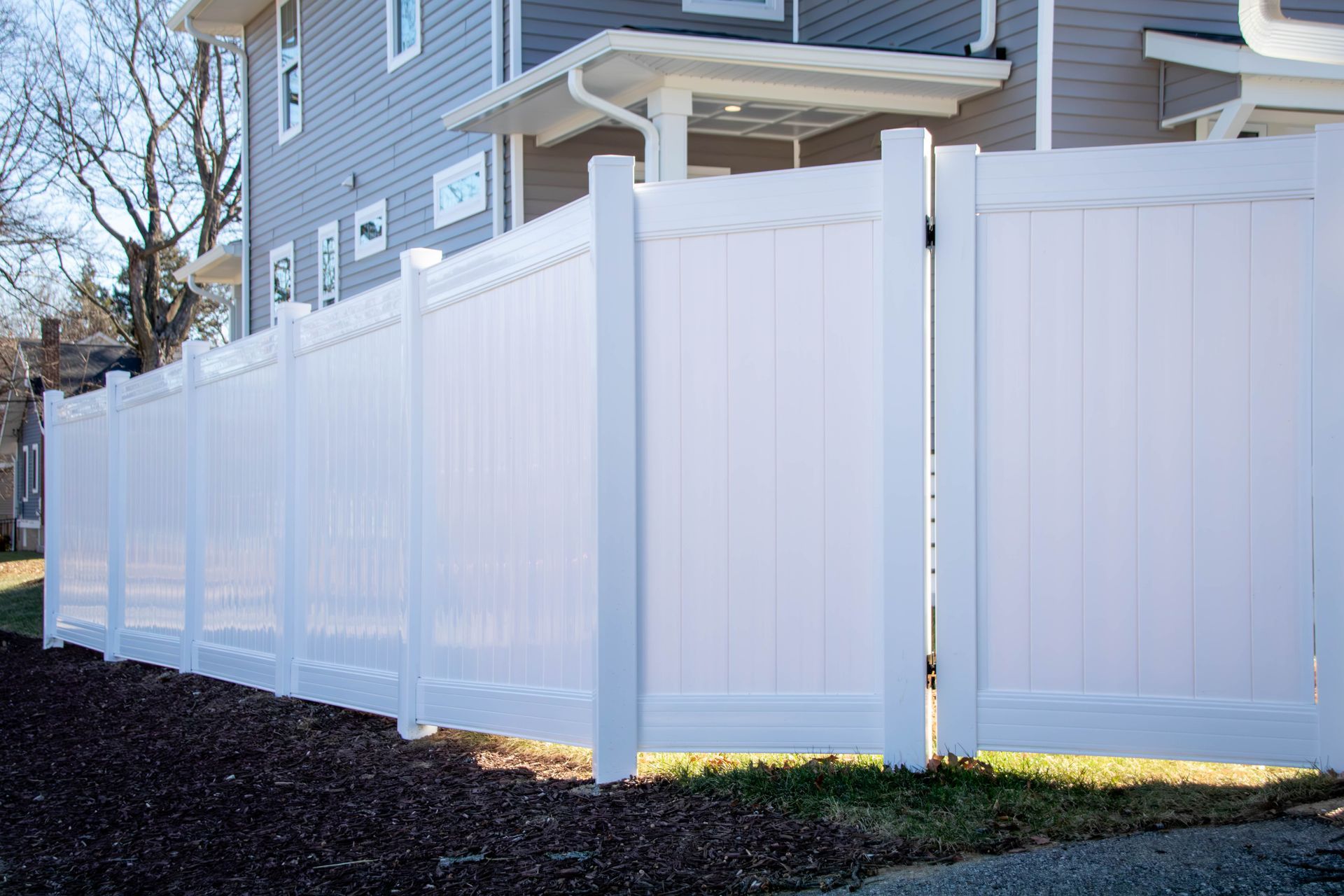 White vinyl privacy fence in front of a gray building, with a gate, on a sunny day.