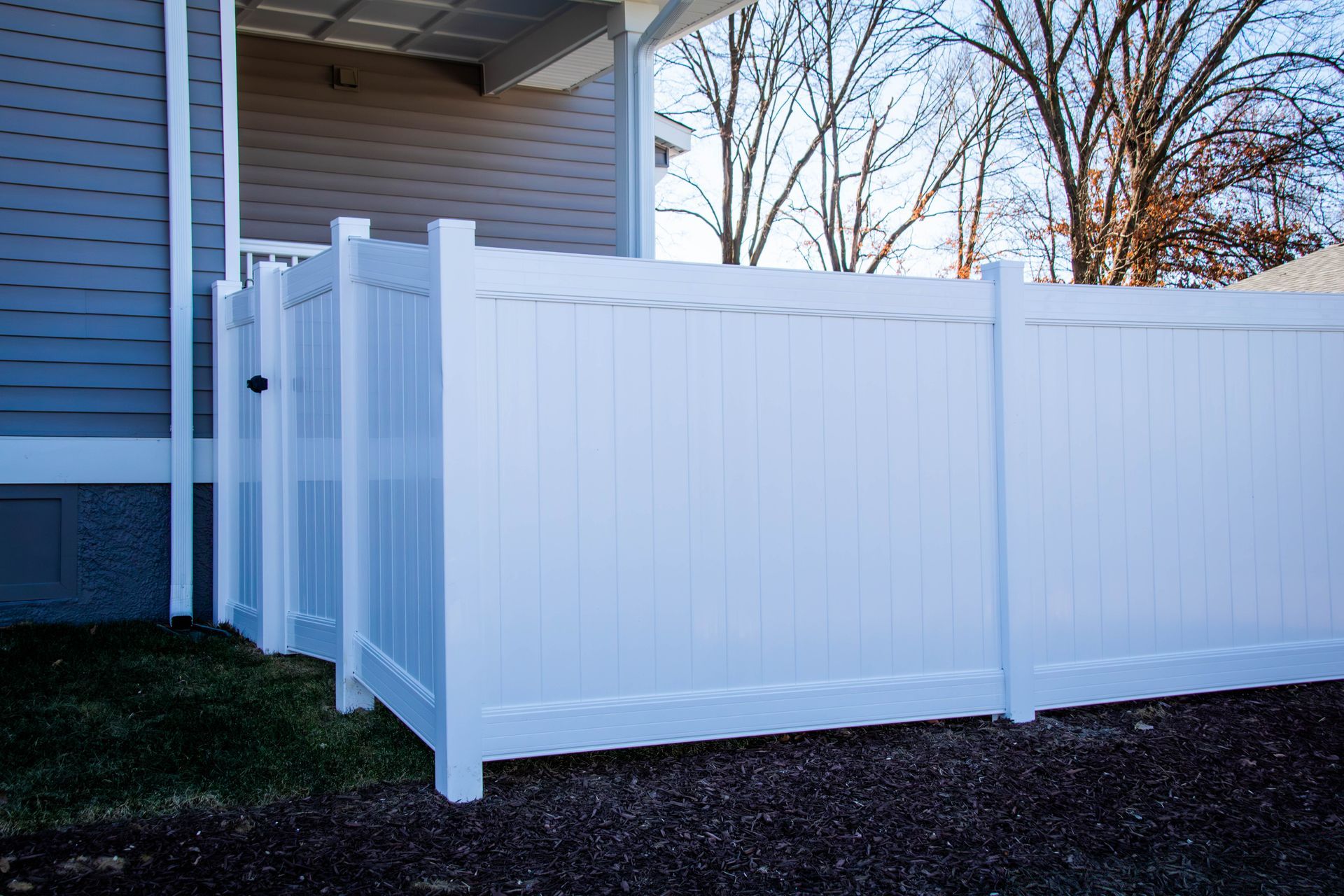 White vinyl fence next to a house, partially obscuring a grassy area and bare trees.