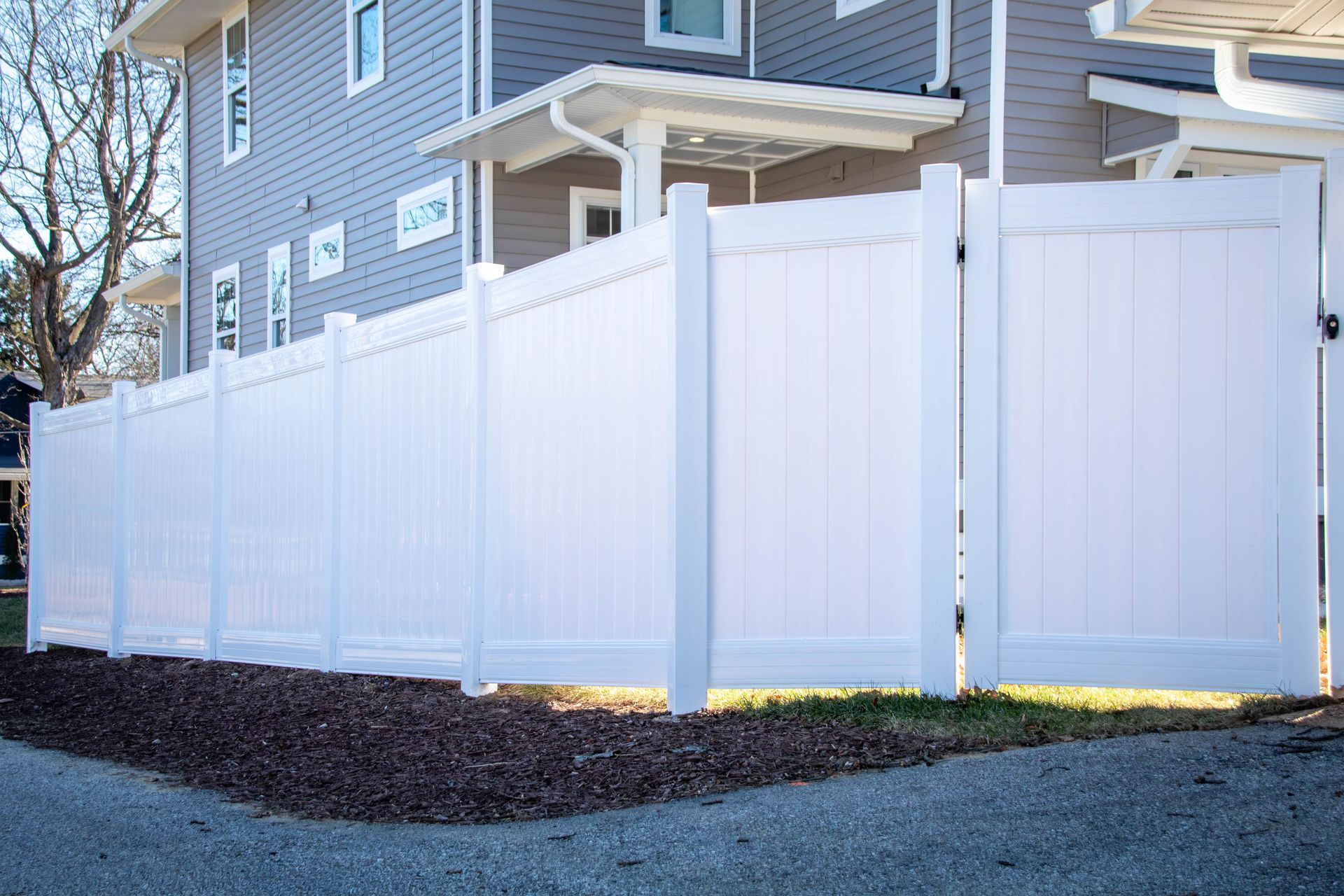 White vinyl fence bordering a building, set on a gravel and grass patch.