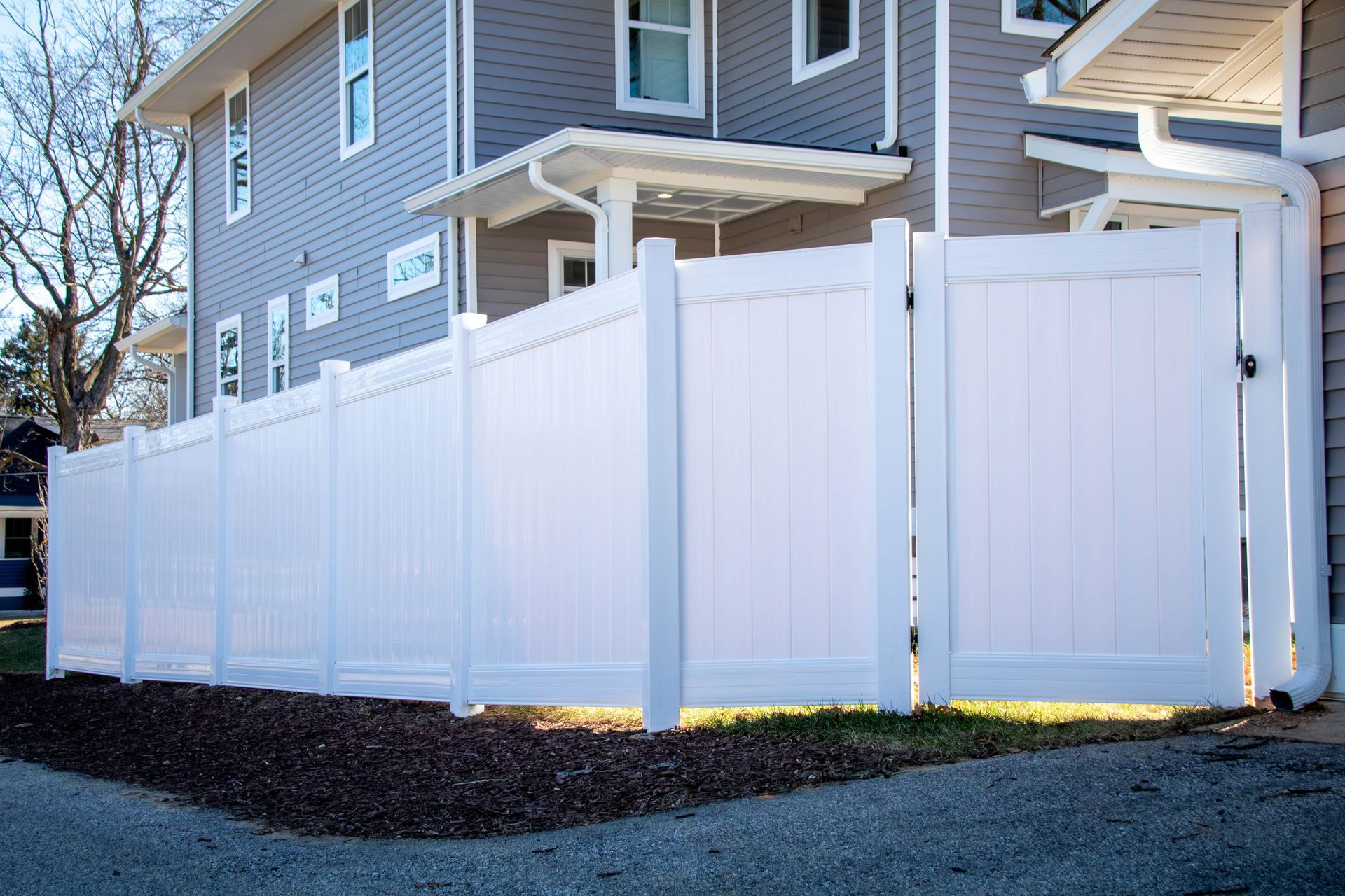 White vinyl fence with gate surrounding a yard next to a gray building.