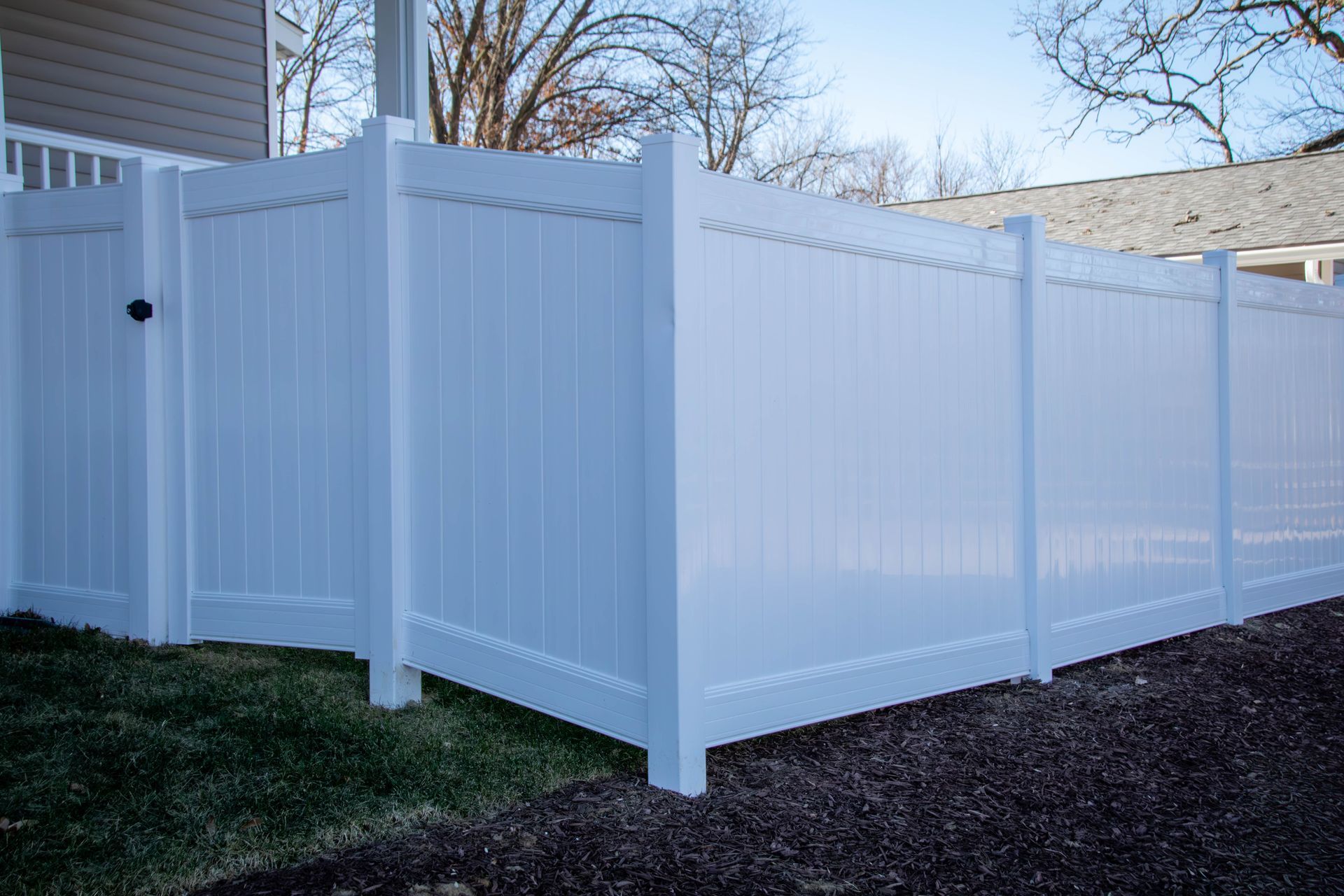 White vinyl fence with gate in front of a house, set on green grass and gravel.
