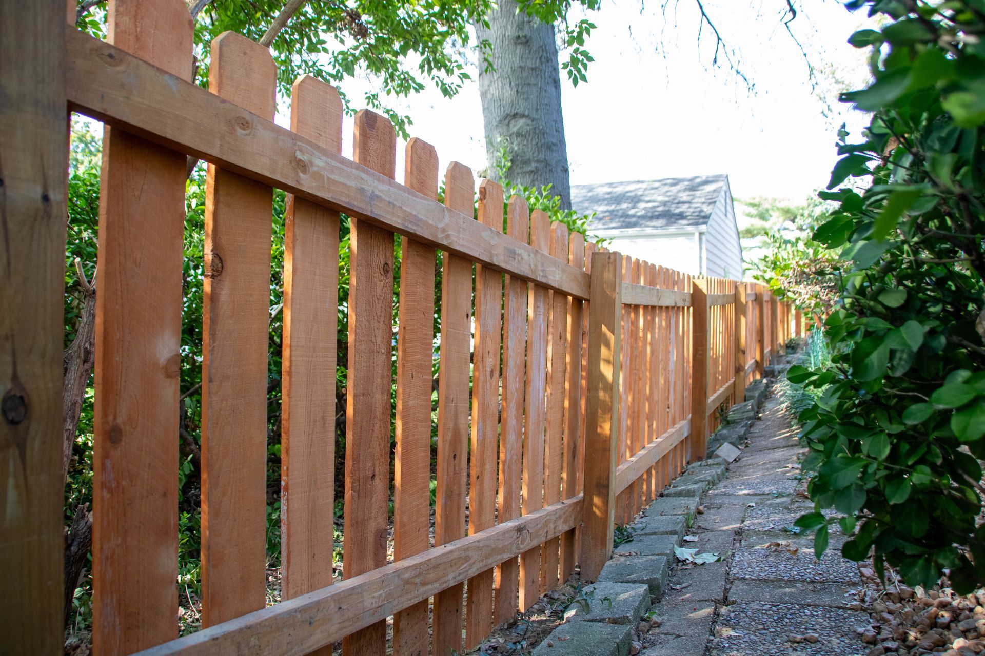 Wooden picket fence bordering a brick pathway, surrounded by greenery and a white house in the distance.