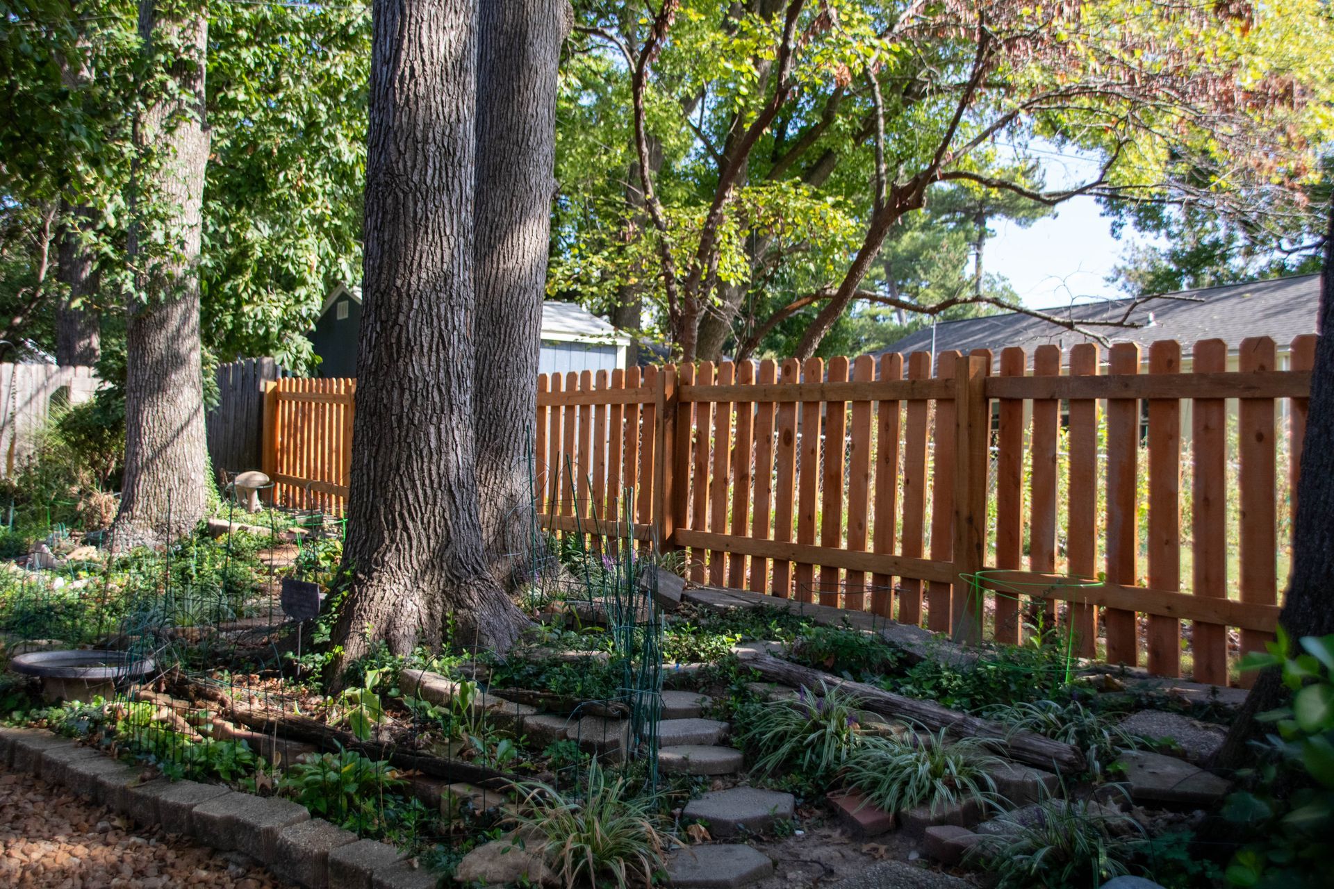 Brown wooden picket fence in a backyard garden with trees and stepping stones.