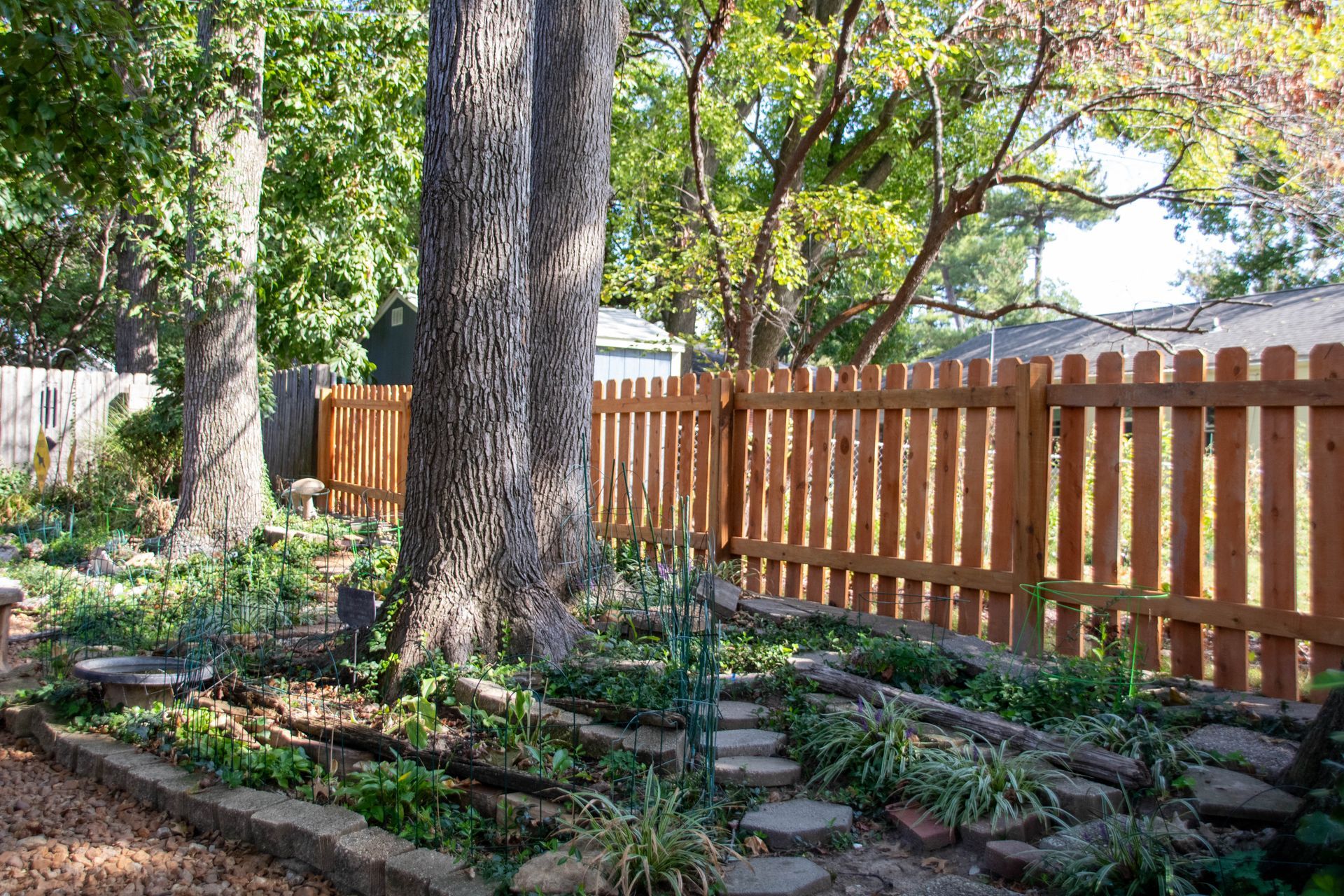 Backyard with trees, garden beds, brown picket fence, and stone pathway.