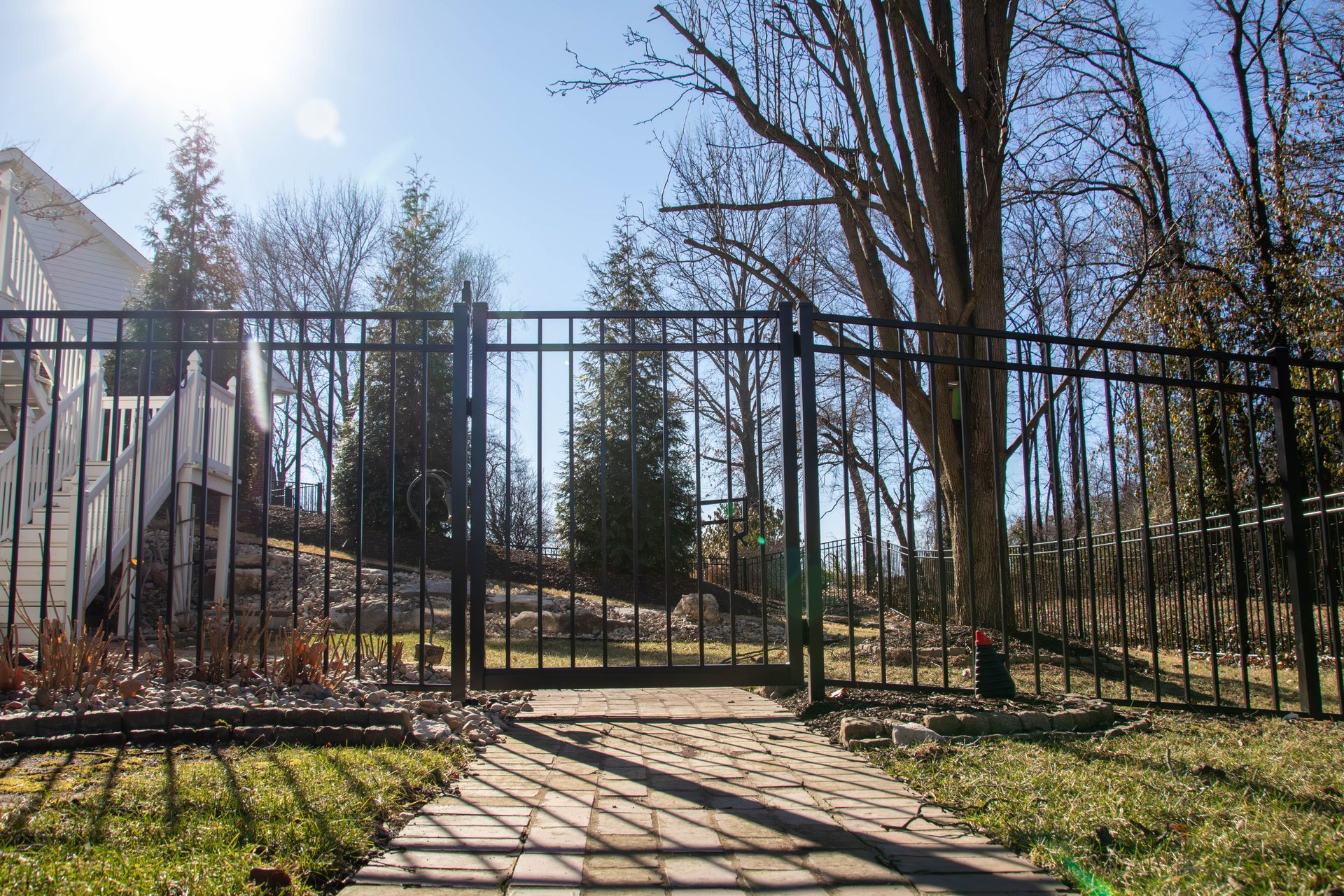 Black metal fence with gate, leading to a yard with trees and a paved path. Sunny day.