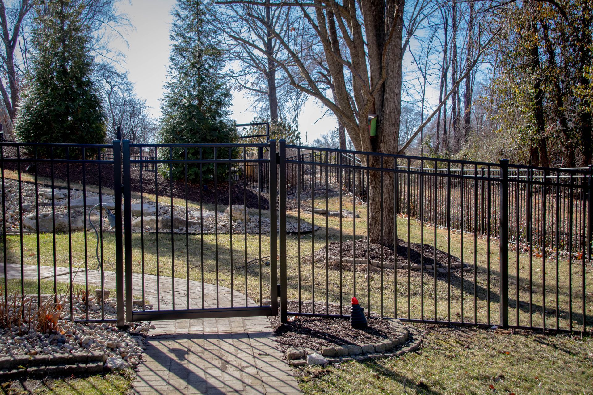 Black metal fence with gate, enclosing a yard with trees and a brick path.