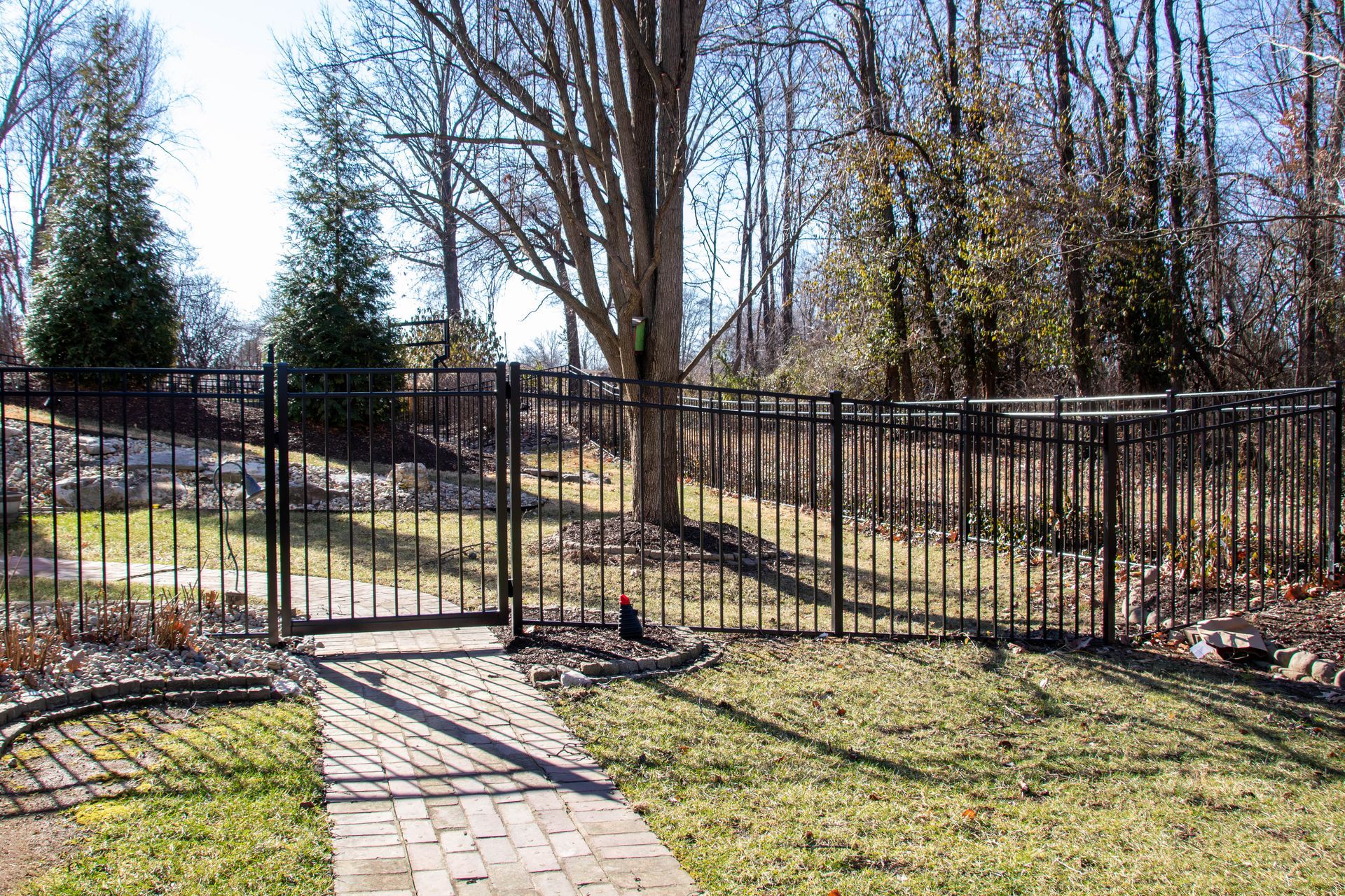 Black metal fence with gate surrounds a brick path and grassy yard near trees.