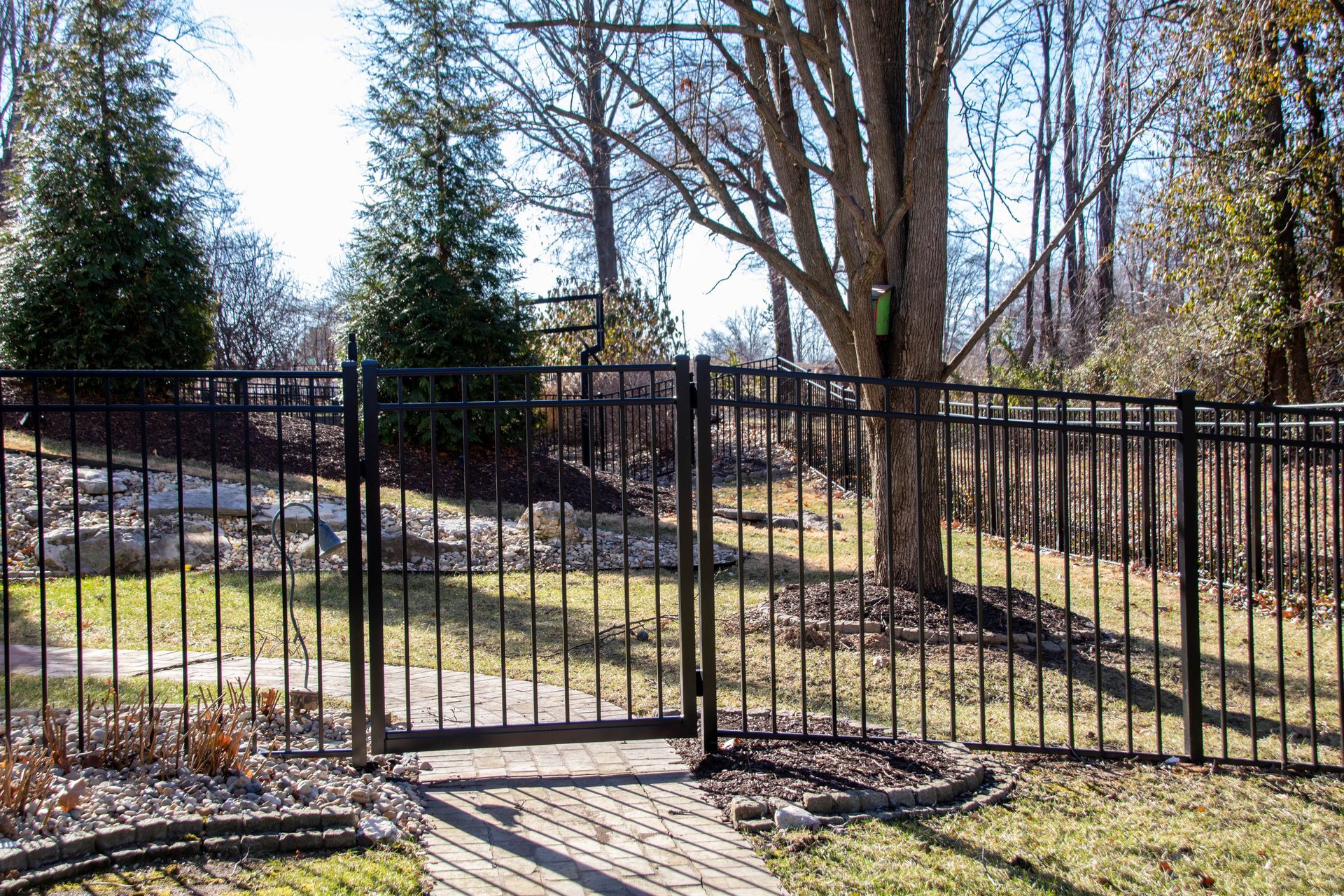 Black metal fence with gate in a yard with grass, trees, and stone pathway.