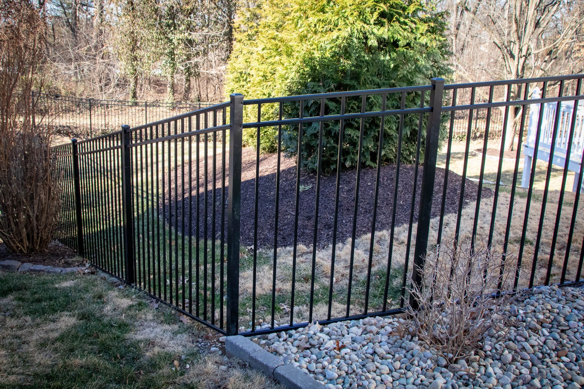 Black metal fence enclosing a garden bed with mulch and a green bush. Sunny outdoor setting.