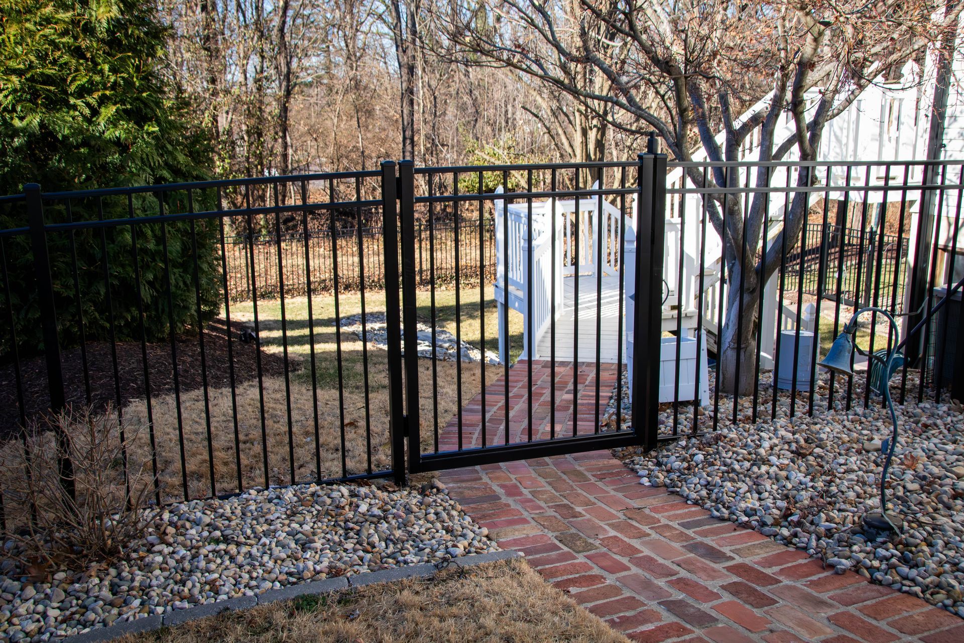 Black metal fence with gate in a backyard. Brick path, gravel, and trees visible.