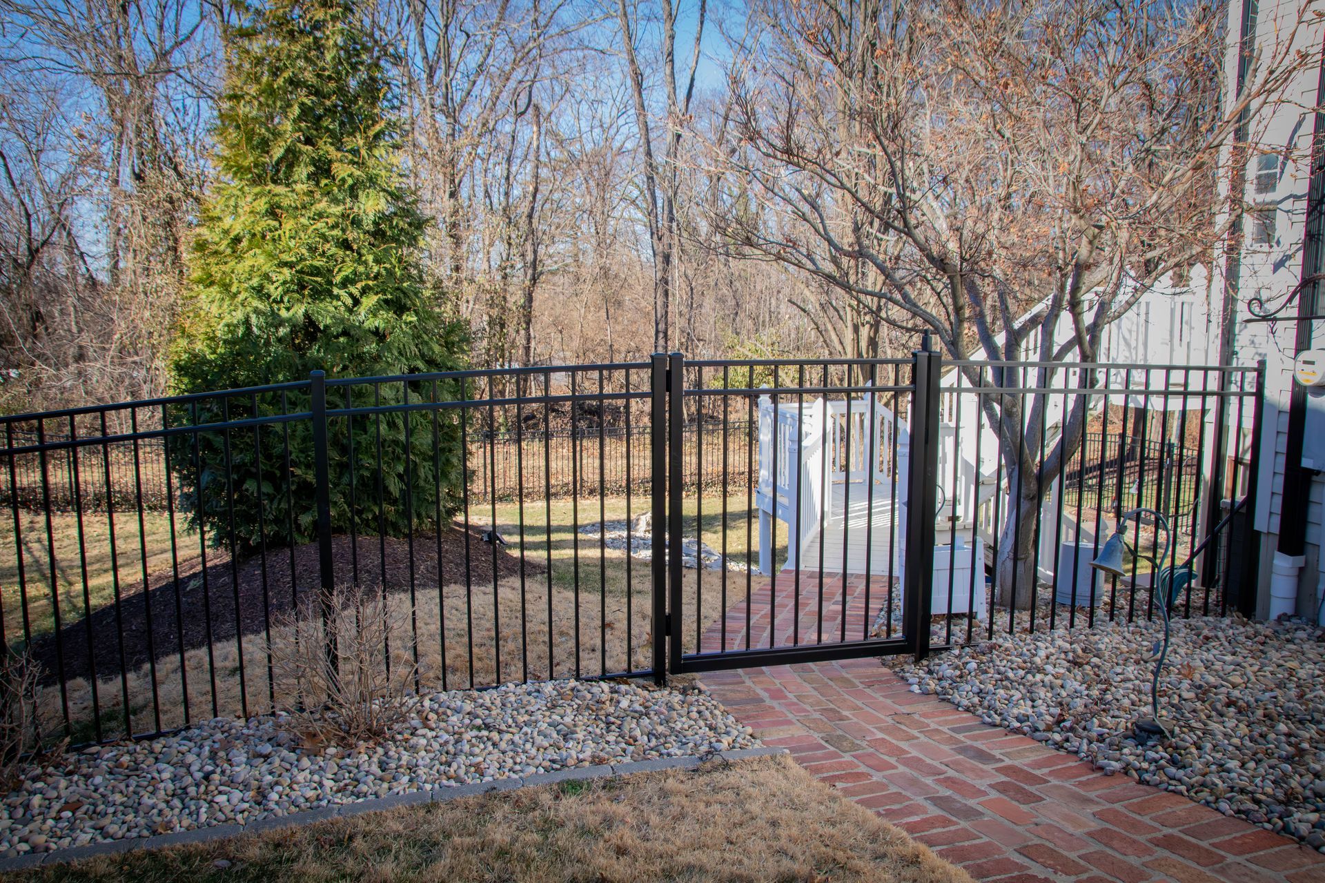 Black metal fence with gate, enclosing a yard with brick path, shrubs, and trees.