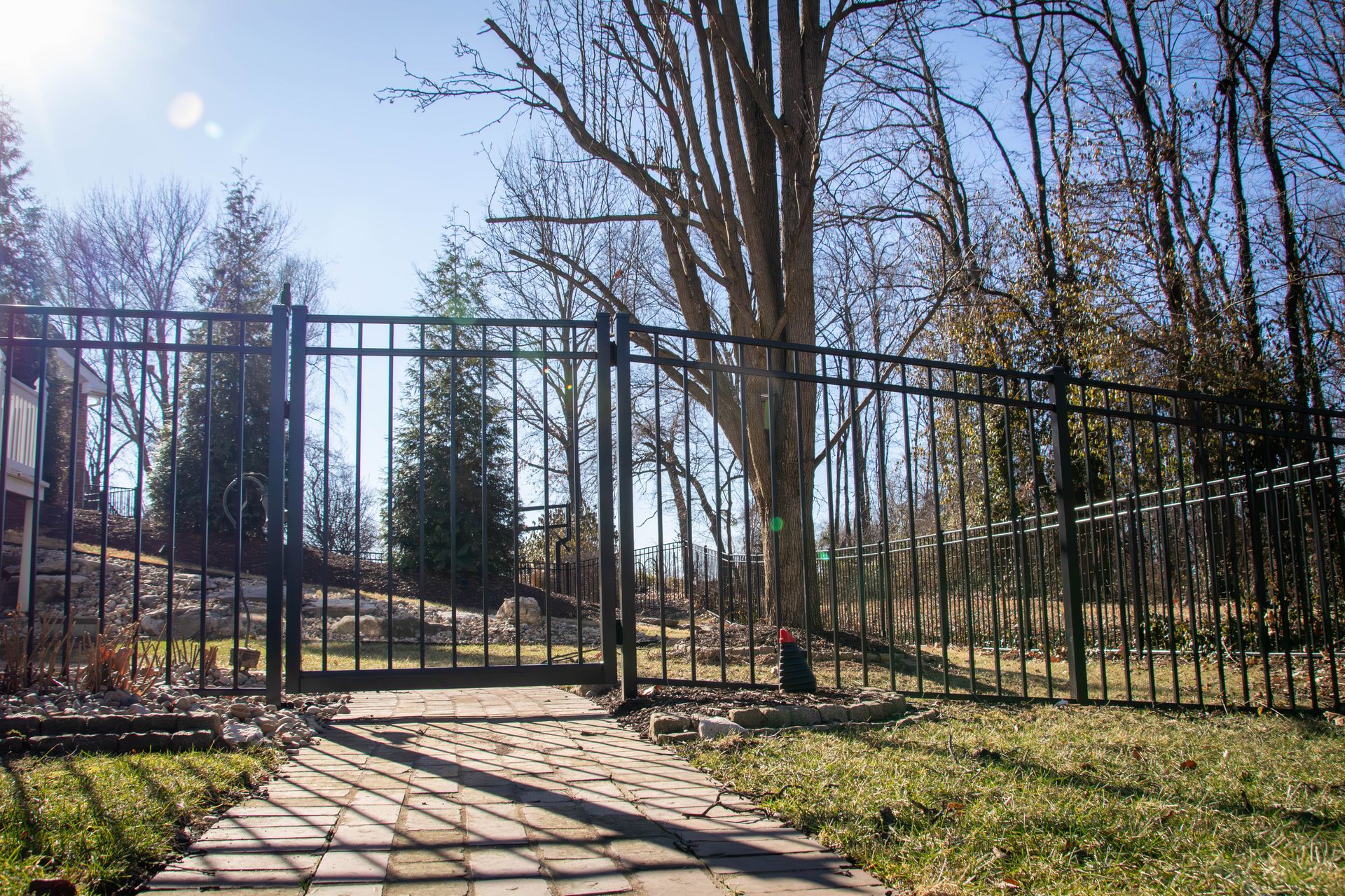 Black metal fence with gate in front of a brick path and grassy yard. Trees and blue sky background.