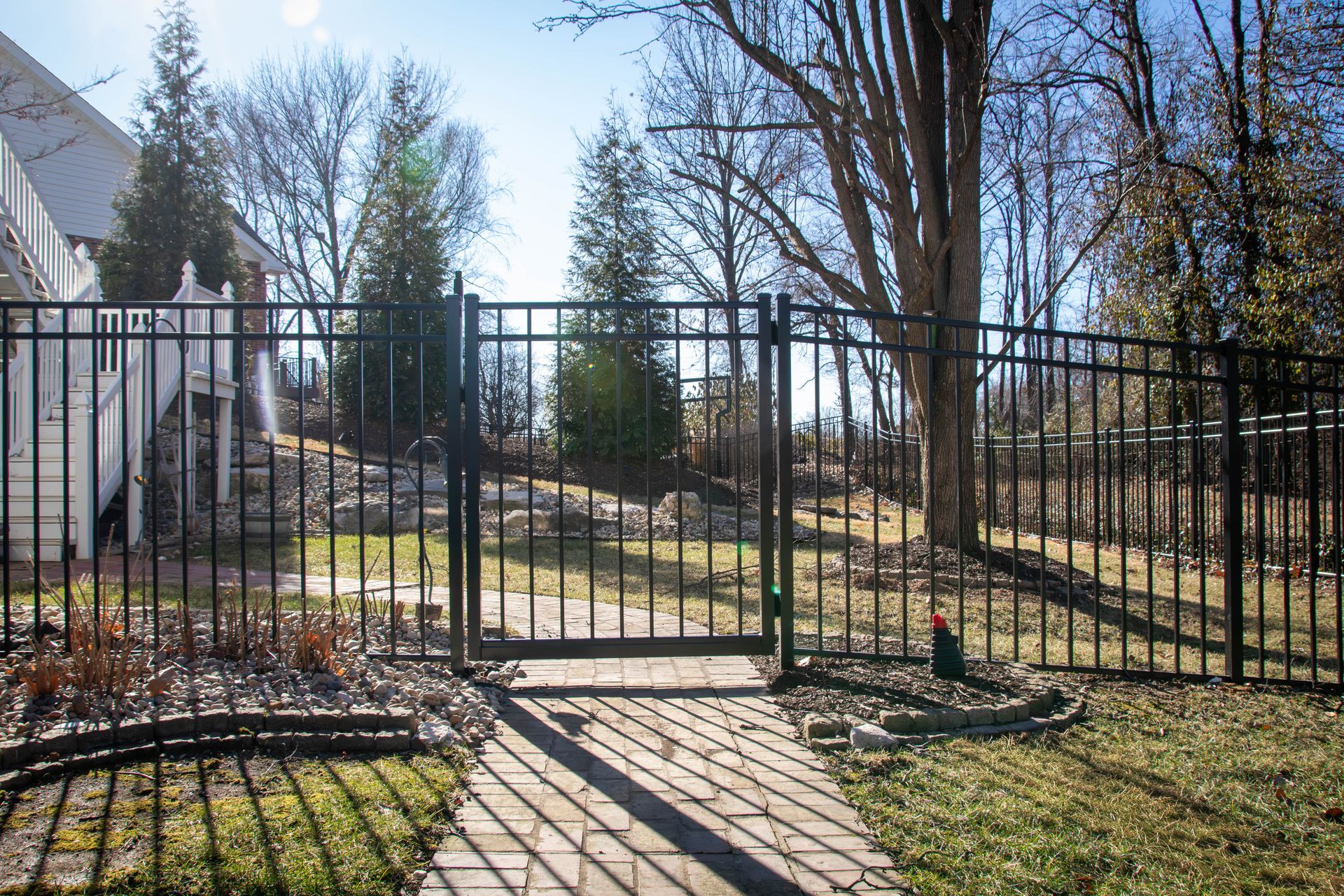 Black metal gate and fence in a backyard, leading to a grassy hill and trees under a blue sky.