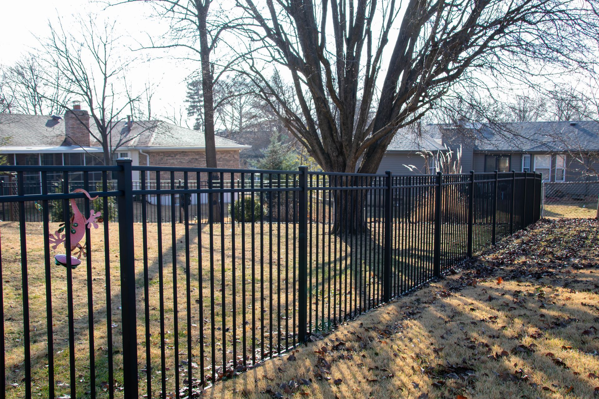 Black metal fence encloses a yard with houses in the background and a tree in the center.