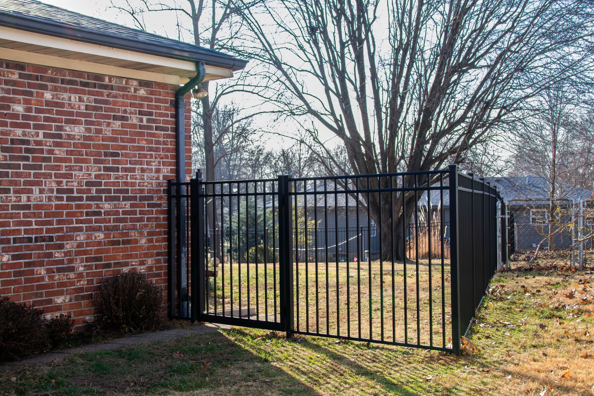 Black metal fence with gate near a brick building and a bare tree.