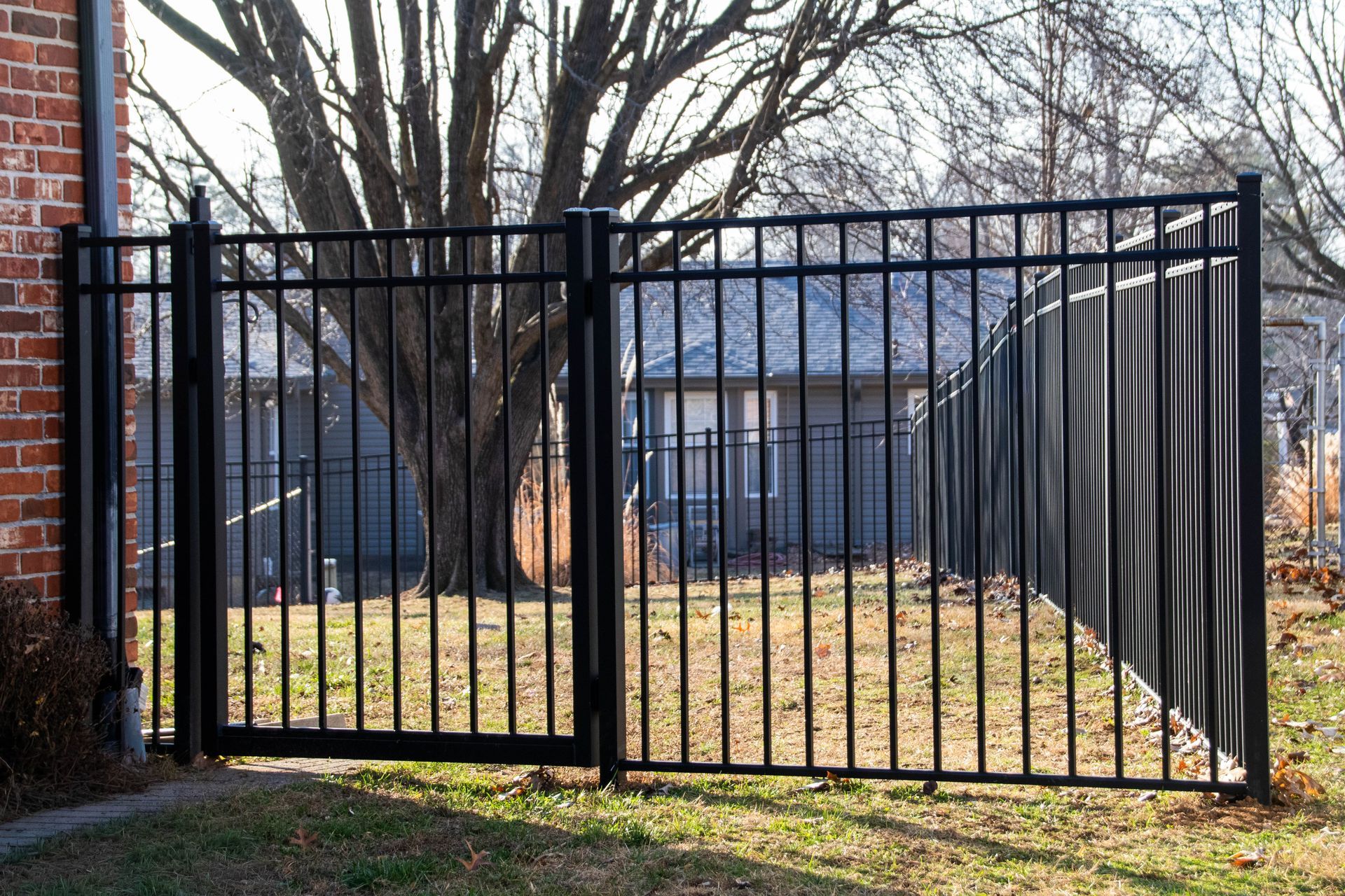 Black metal fence with a gate, in front of a house, and next to a brick building.