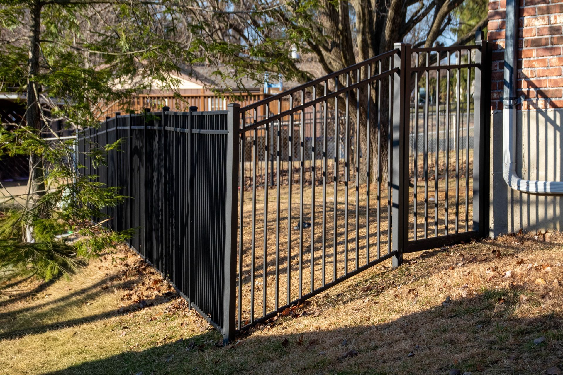 Black metal fence with gate, bordering a yard with brown grass and a brick wall.