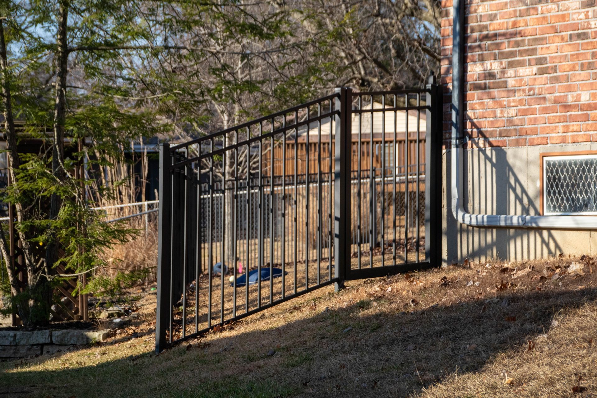Black metal fence next to a brick building on a sloped grassy yard, with trees in the background.