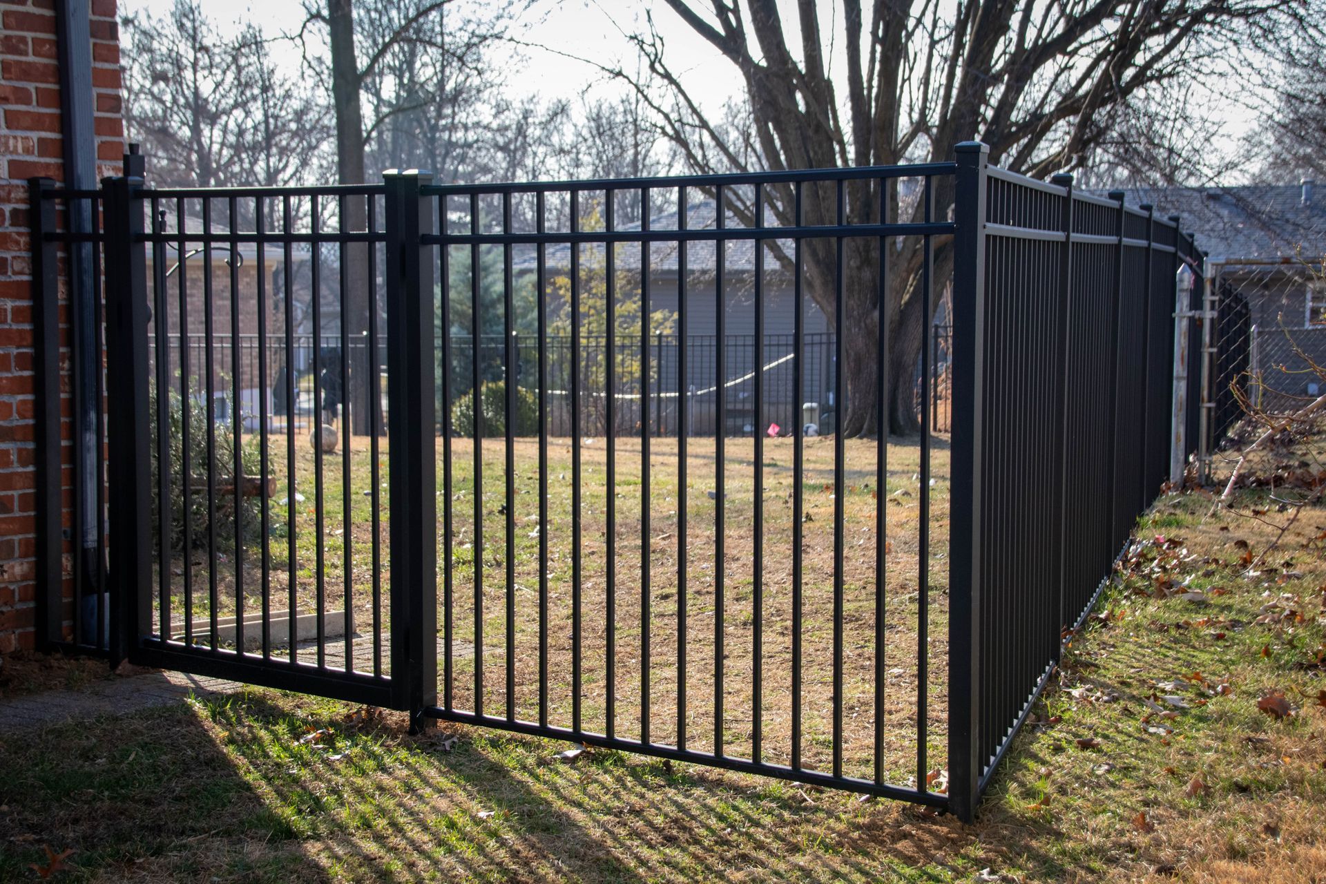 Black metal fence surrounding a yard with bare grass, brick building, and trees in background.