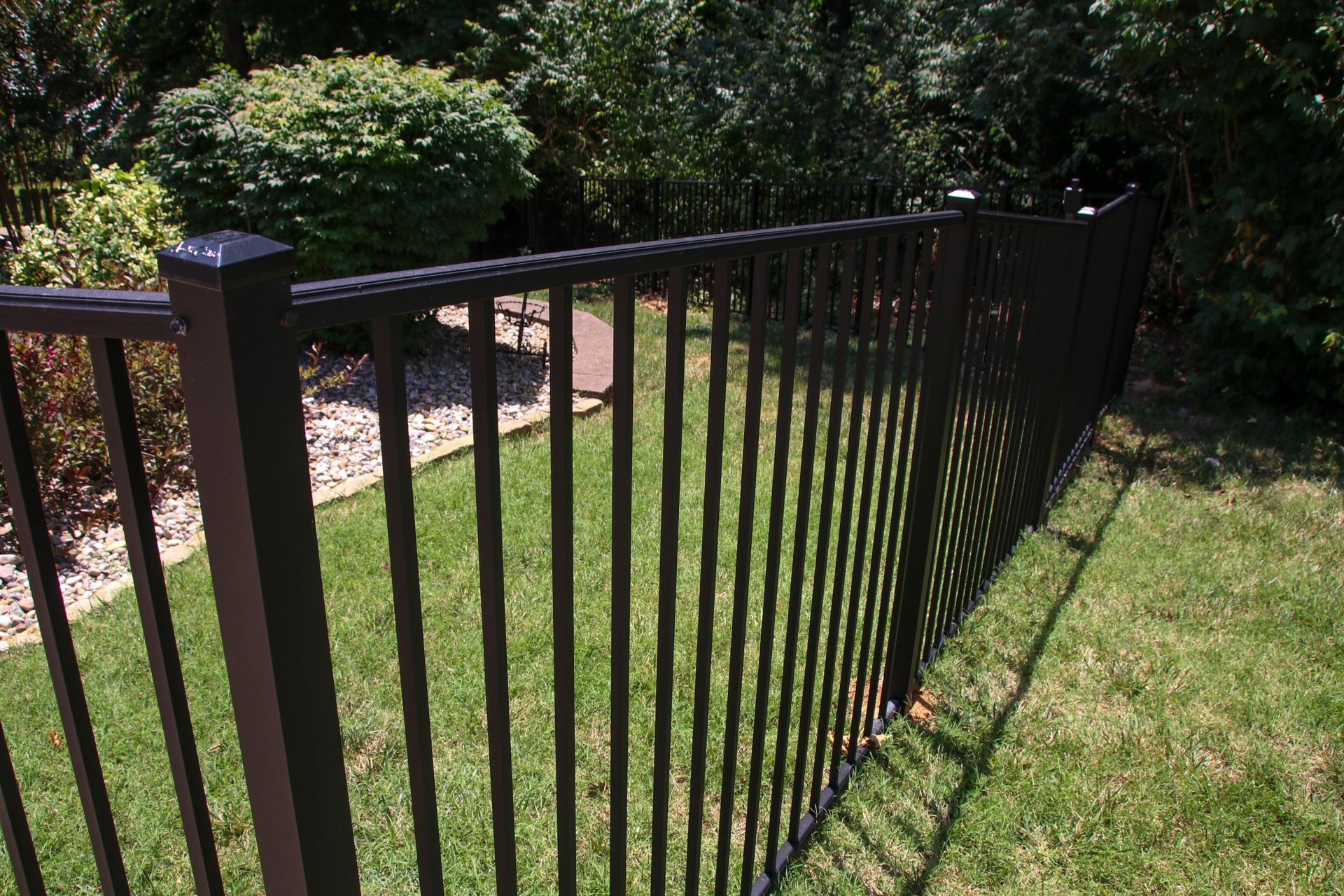 Black metal fence bordering a green lawn, with shrubs and trees in the background.