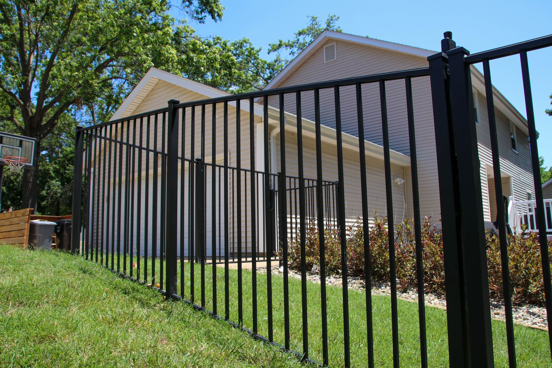 Black metal fence in front of a tan house on a sunny day. Green grass and shrubs.