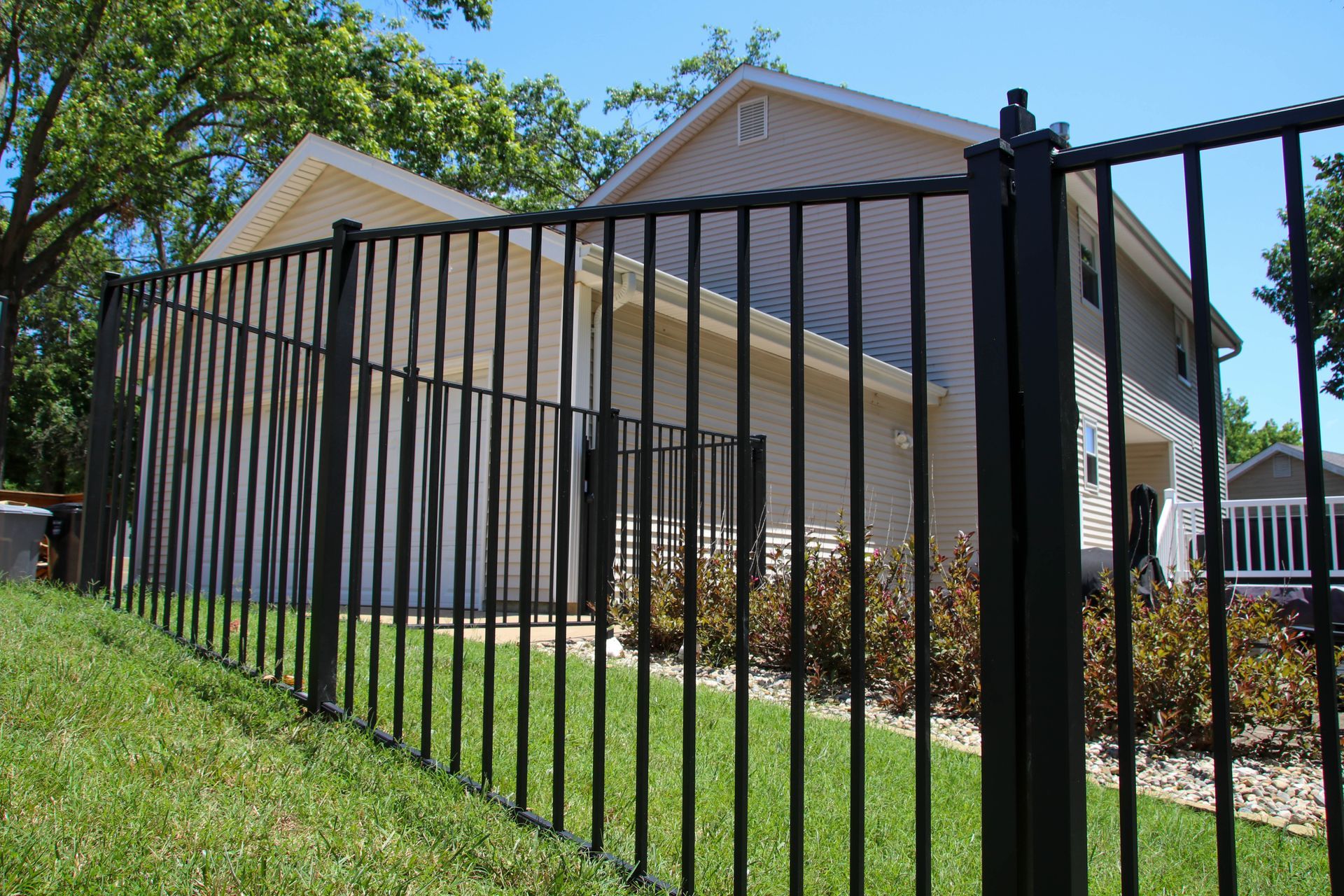 Black metal fence surrounding a two-story beige house with a green lawn under a bright blue sky.