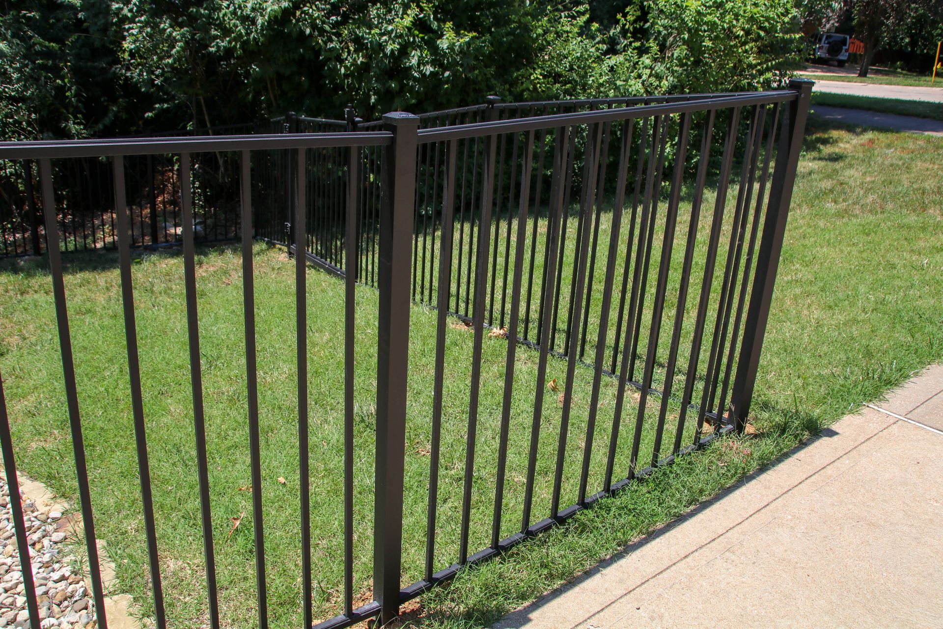 Dark metal fence enclosing a grassy yard next to a sidewalk and bushes.
