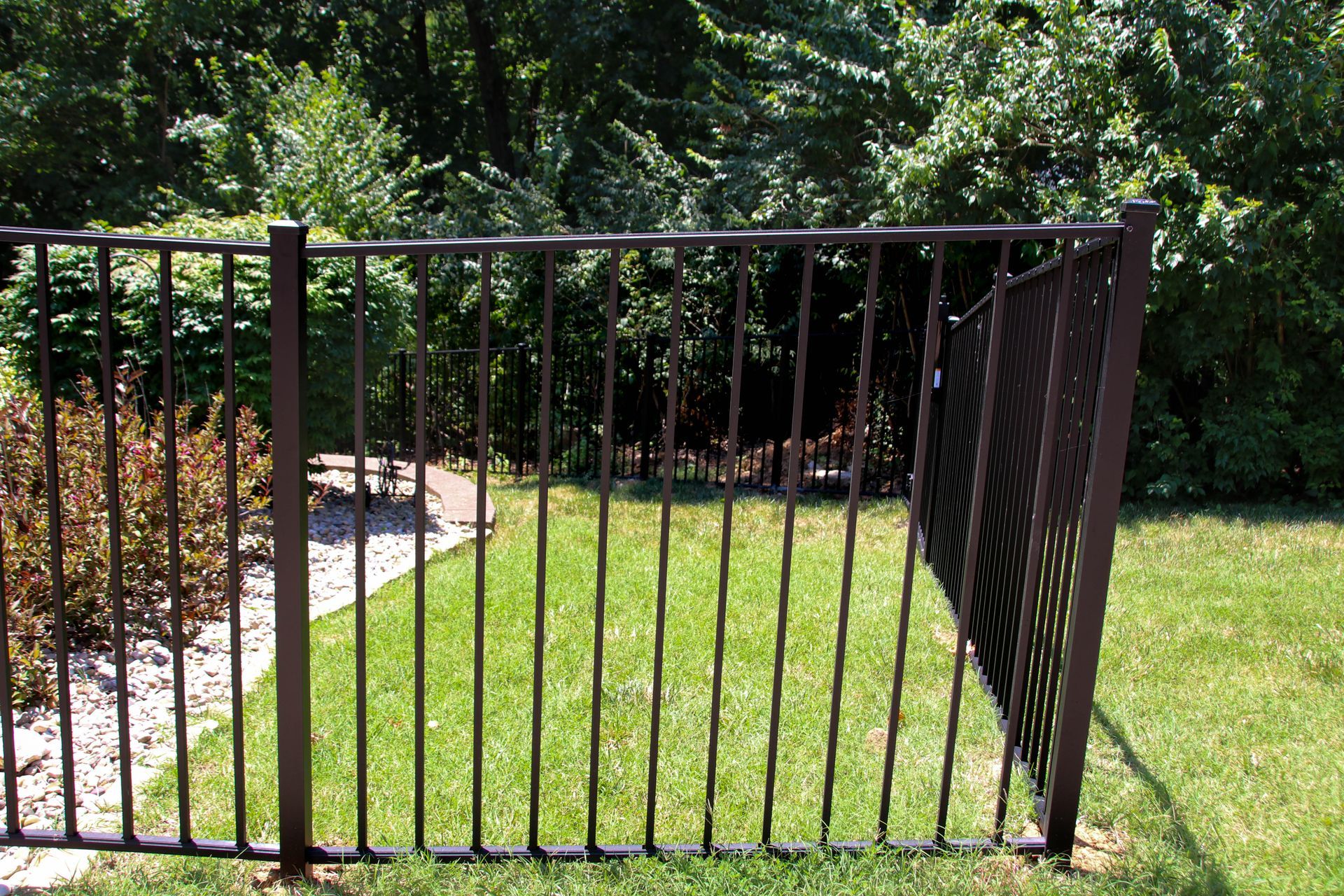 Brown metal fence enclosing a grassy yard, with trees in the background.