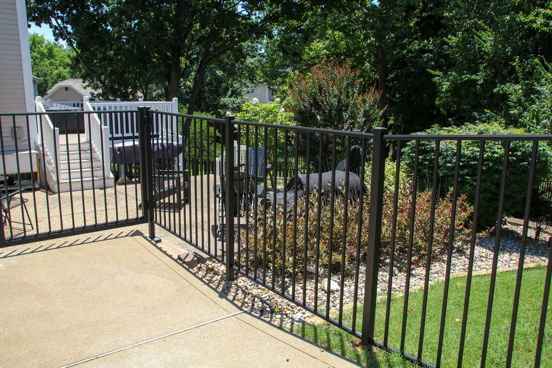 Black metal fence surrounding a concrete patio and a grassy area, with a gate, trees, and house in the background.