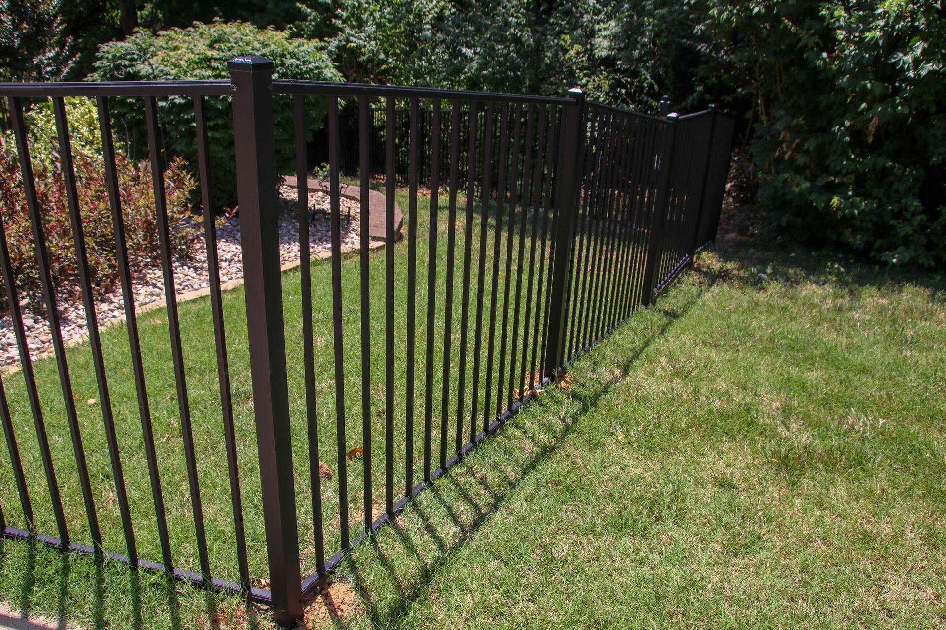 Black metal fence on green grass, separating yard from trees and bushes.