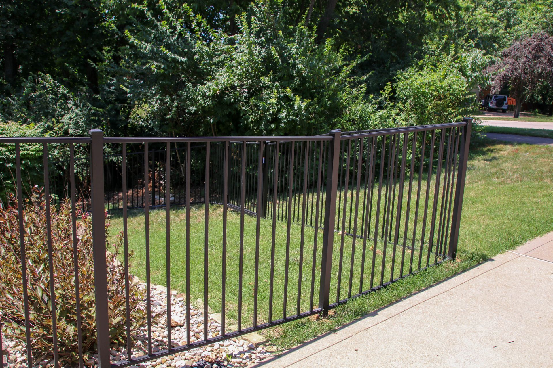 Brown metal fence in front of green grass and bushes, sidewalk on the right.
