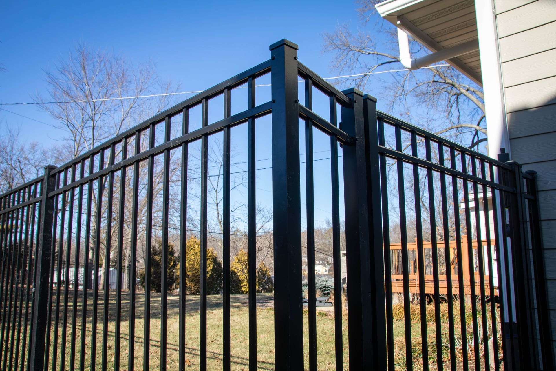 Black metal fence corner next to a house, with a grassy yard and bare trees visible beyond.
