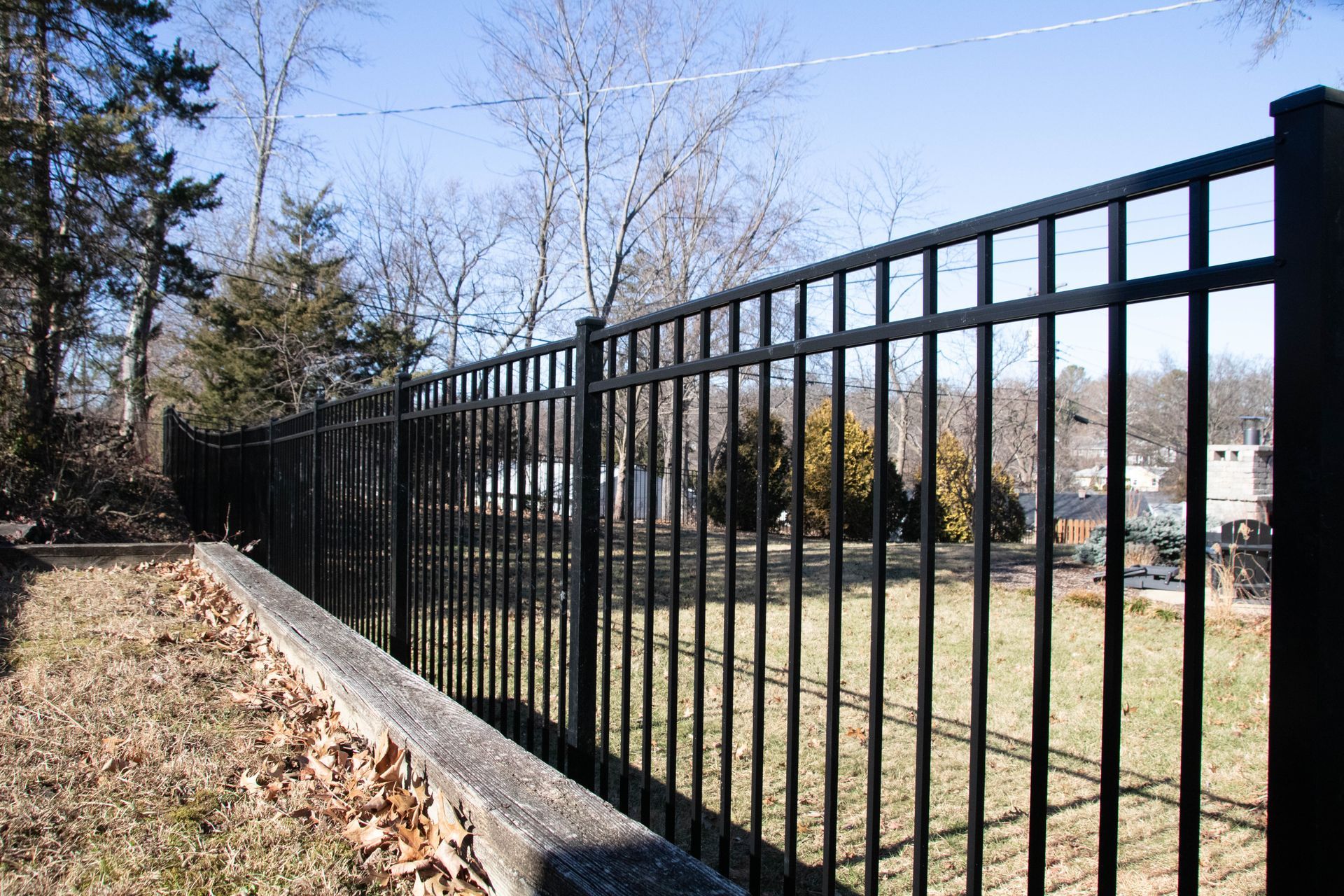 Black metal fence along a stone wall in a grassy area with trees and houses in the background.