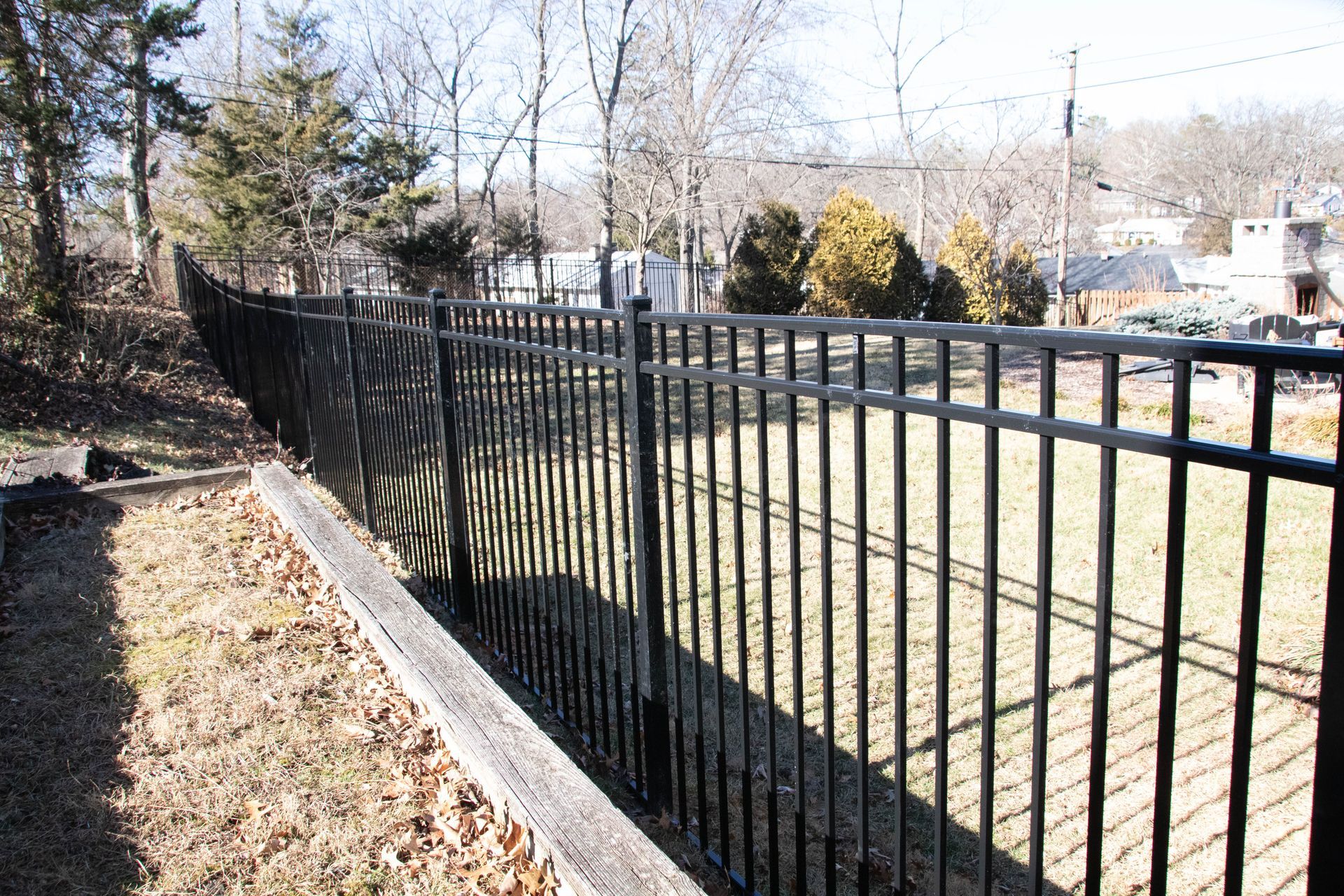 Black metal fence bordering dry grass and trees in sunlight.