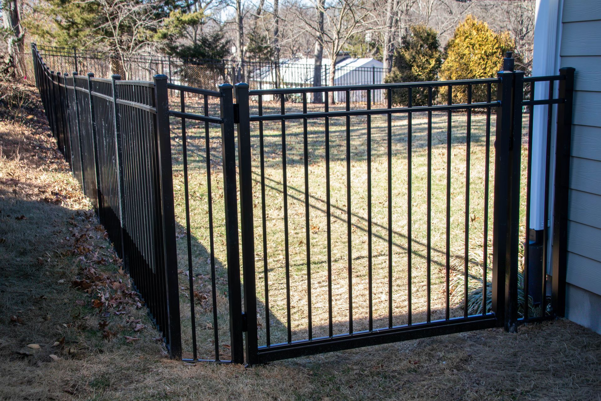 Black metal fence enclosing a grassy yard.  A house wall is on the right.  A privacy screen is attached to part of the fence.