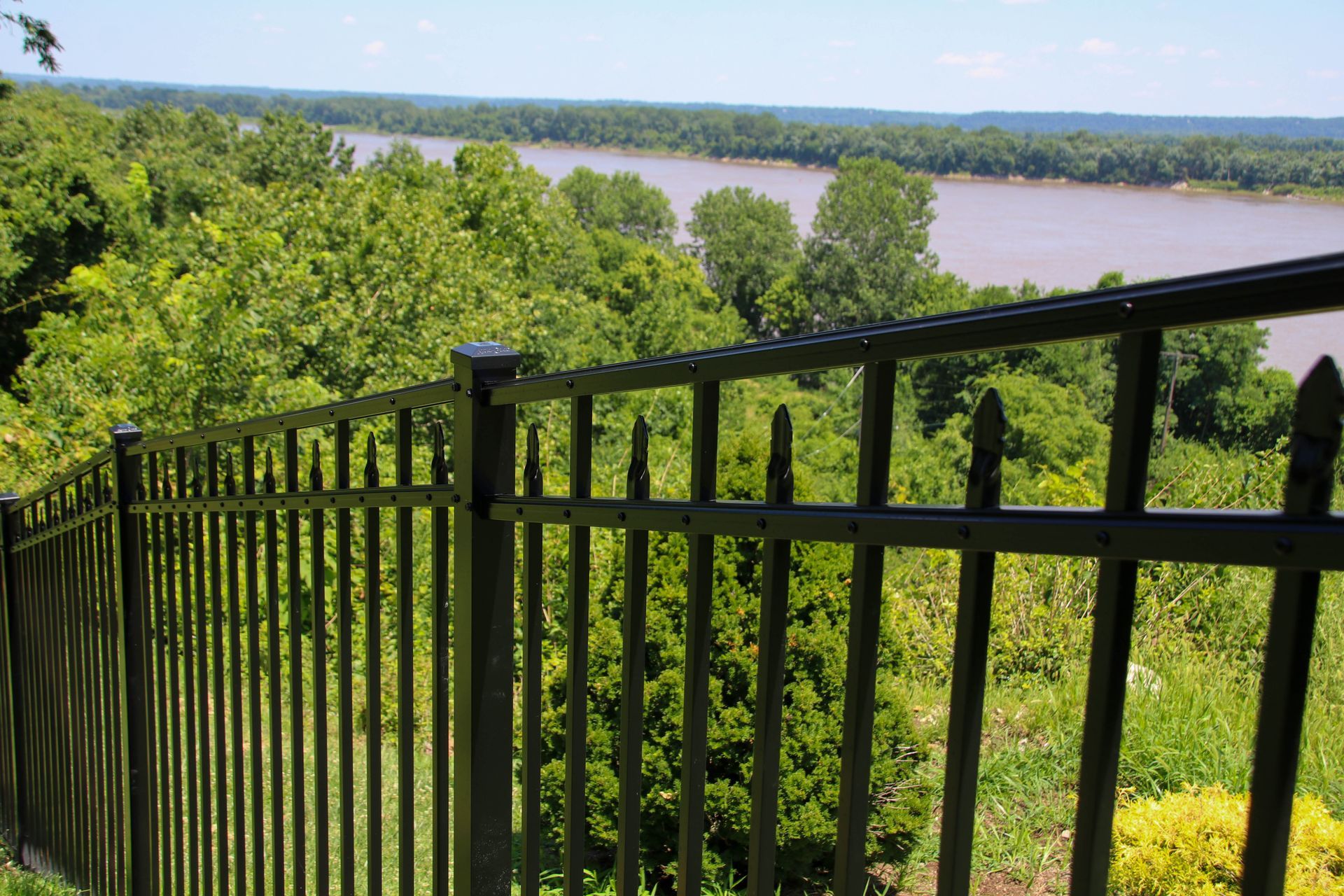 Black metal fence overlooking a river and lush green trees on a sunny day.