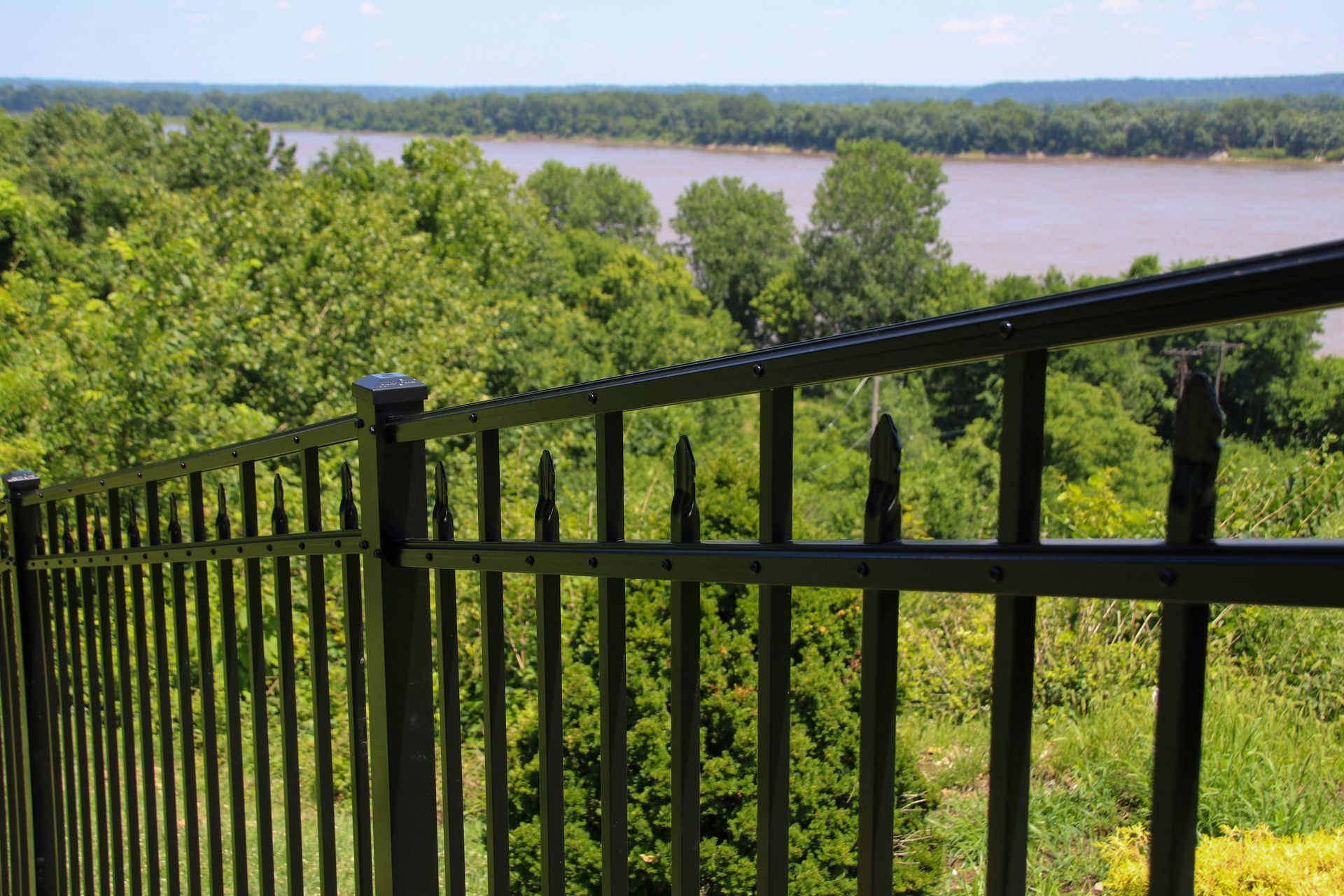Black metal fence overlooking a wide river and green treeline under a blue sky.